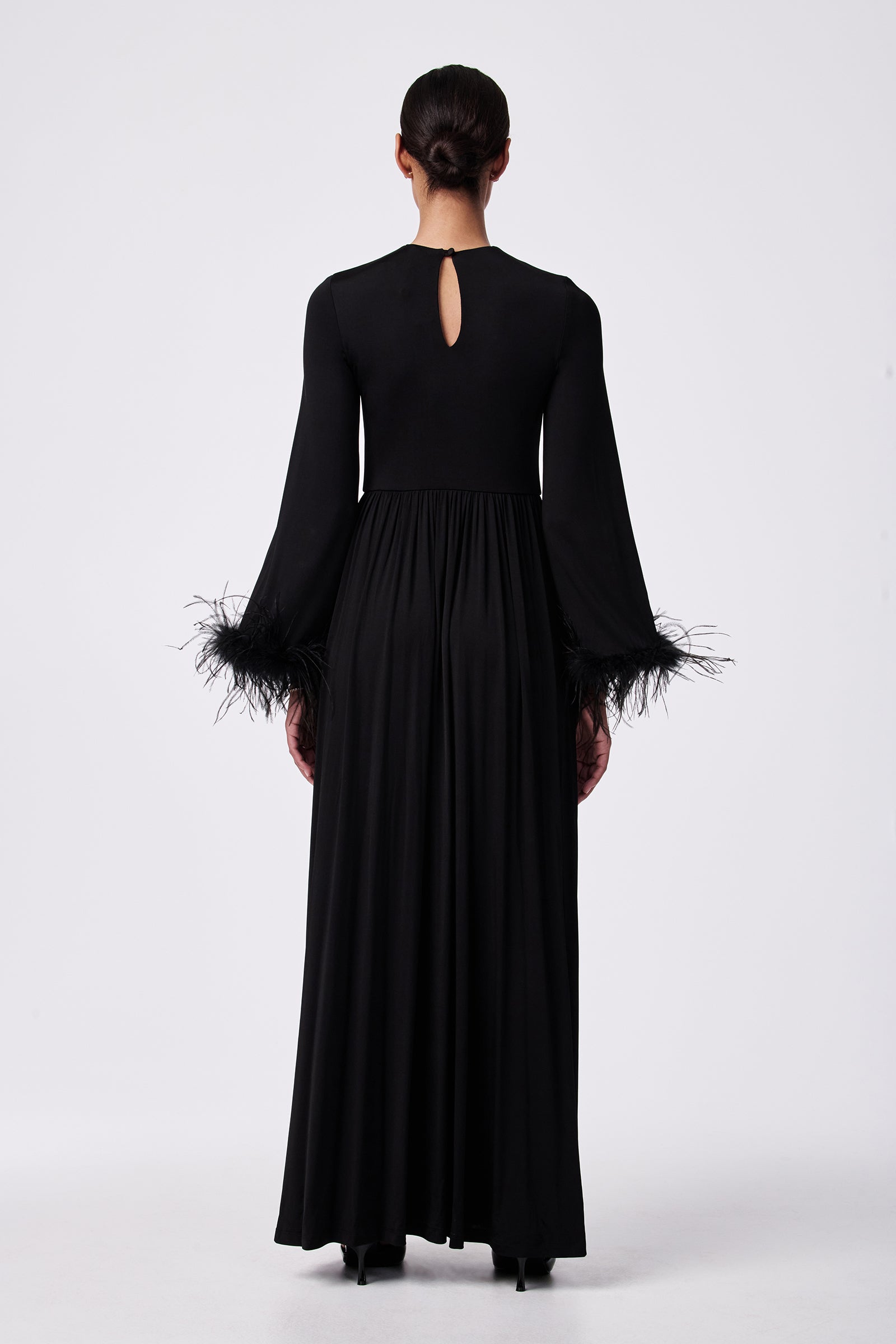 A person stands facing away, wearing the Orla Feather-Cuff Maxi Dress—a long black gown with flared feather-trimmed sleeves, a small keyhole opening at the upper back, and hair styled in a low bun.