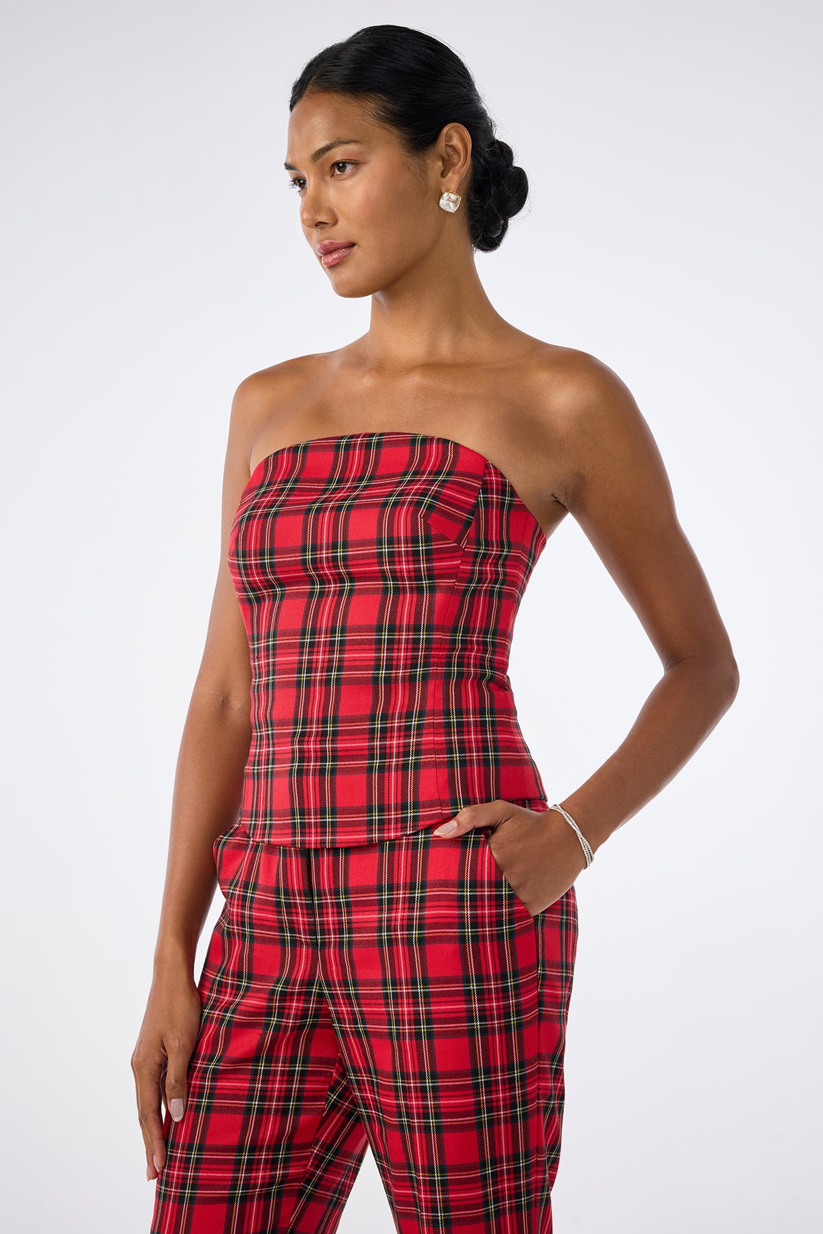 A woman wears the Arden Top, a red plaid strapless piece, paired with matching pants. She stands with one hand in her pocket, looking to the side, her hair in a low bun, accessorized with earrings and a bracelet.