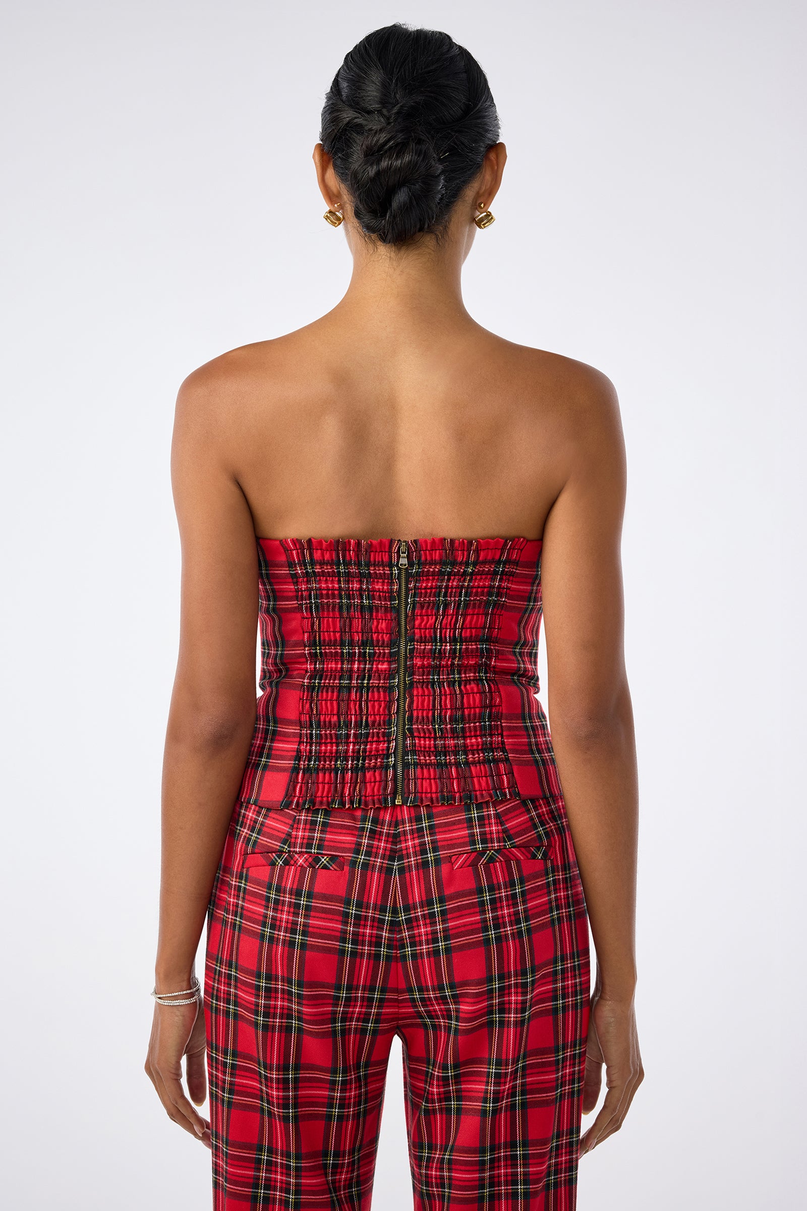 A person is seen from behind wearing the strapless Arden Top in red plaid with matching pants. Their hair is styled in a neat bun, accessorized with bracelets on the left wrist and small hoop earrings against a plain white background.