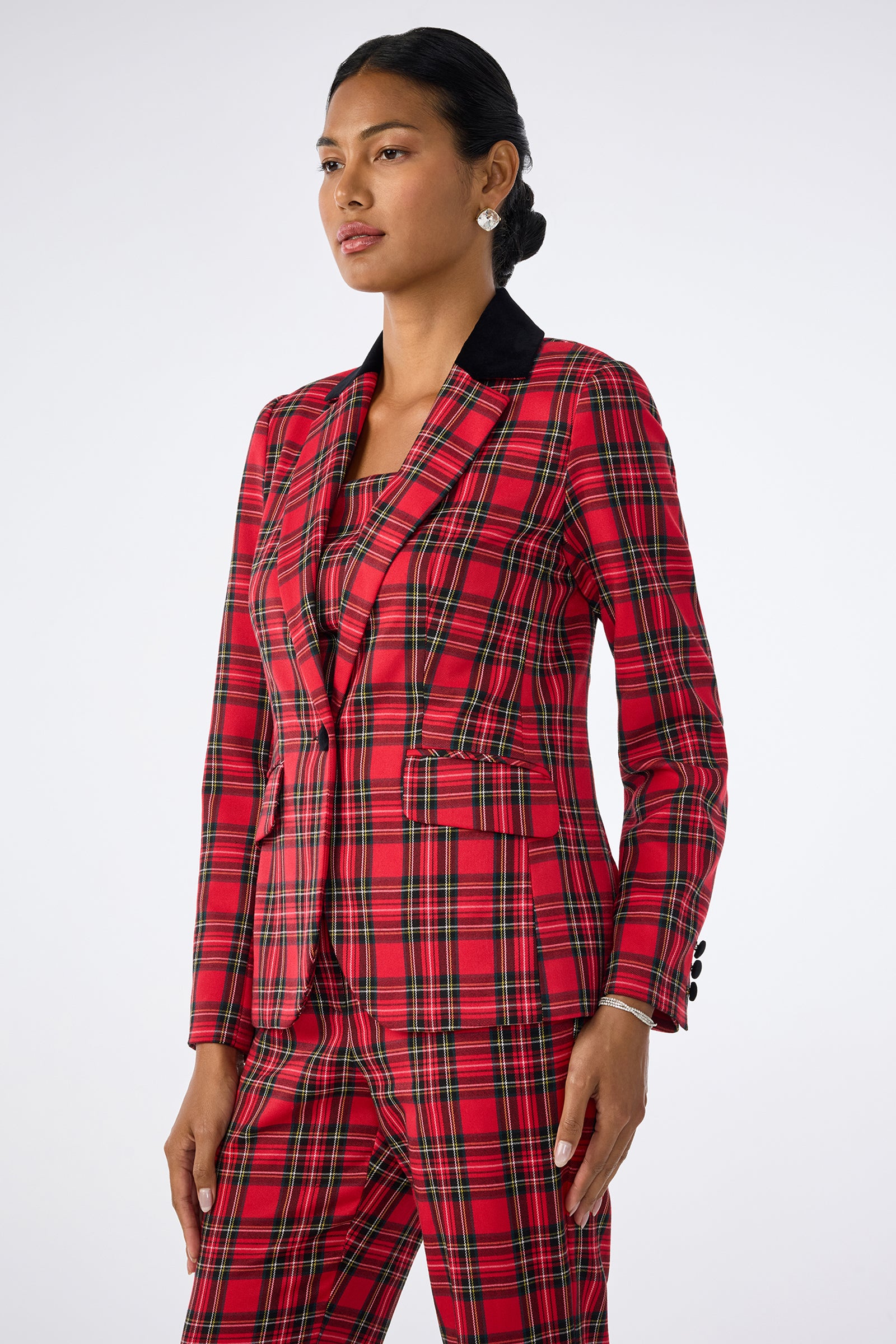 A woman stands confidently against a plain background wearing the Revolution Jacket, a red, black, and yellow plaid blazer with matching pants. Her dark hair is tied back, she wears pearl earrings, and exudes poise.