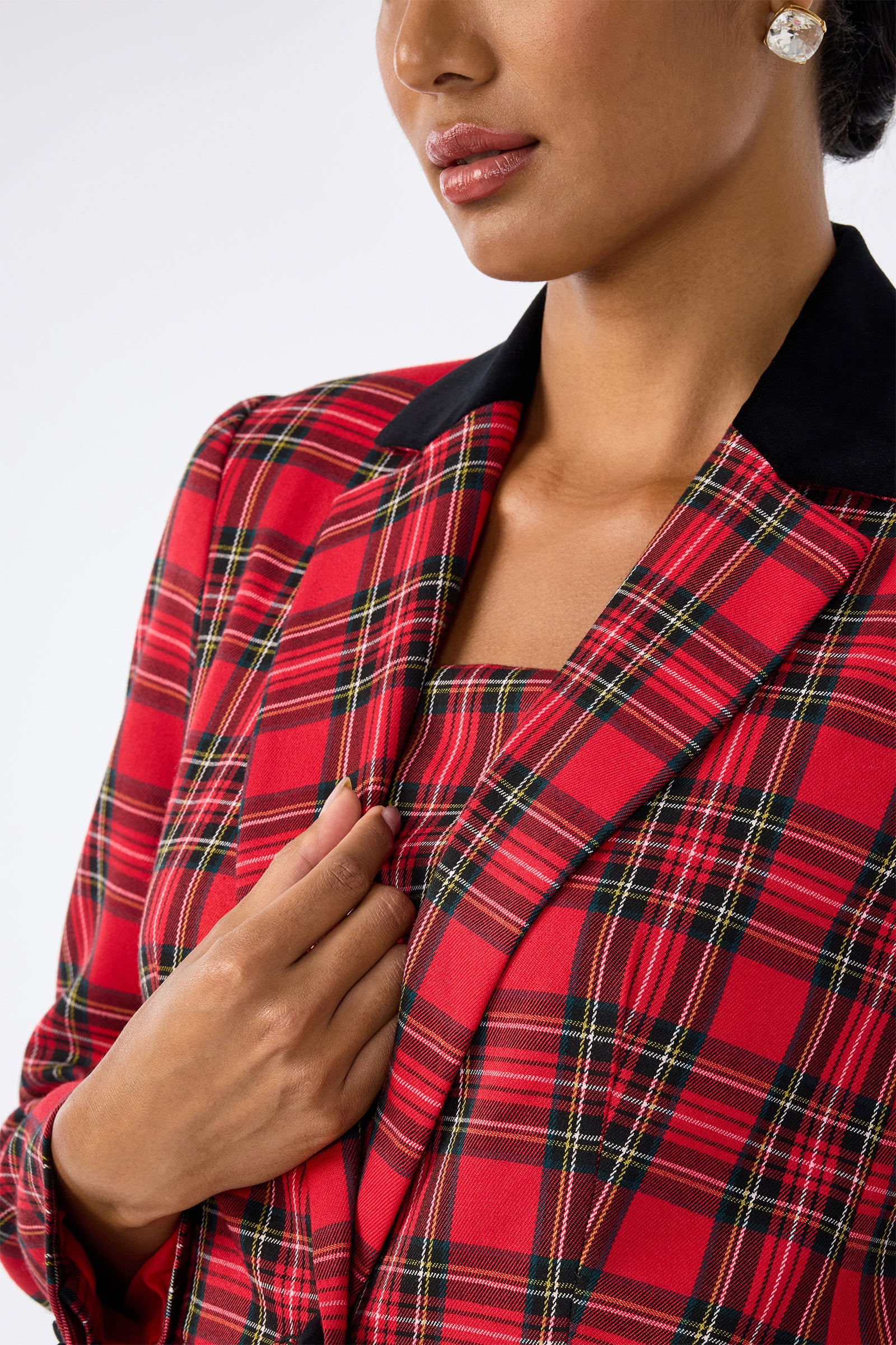 A woman wearing the Revolution Jacket—a red plaid blazer with black lapels and matching top—holds the lapel. Only her lower face and upper body are visible, and she accessorizes with pearl earrings.