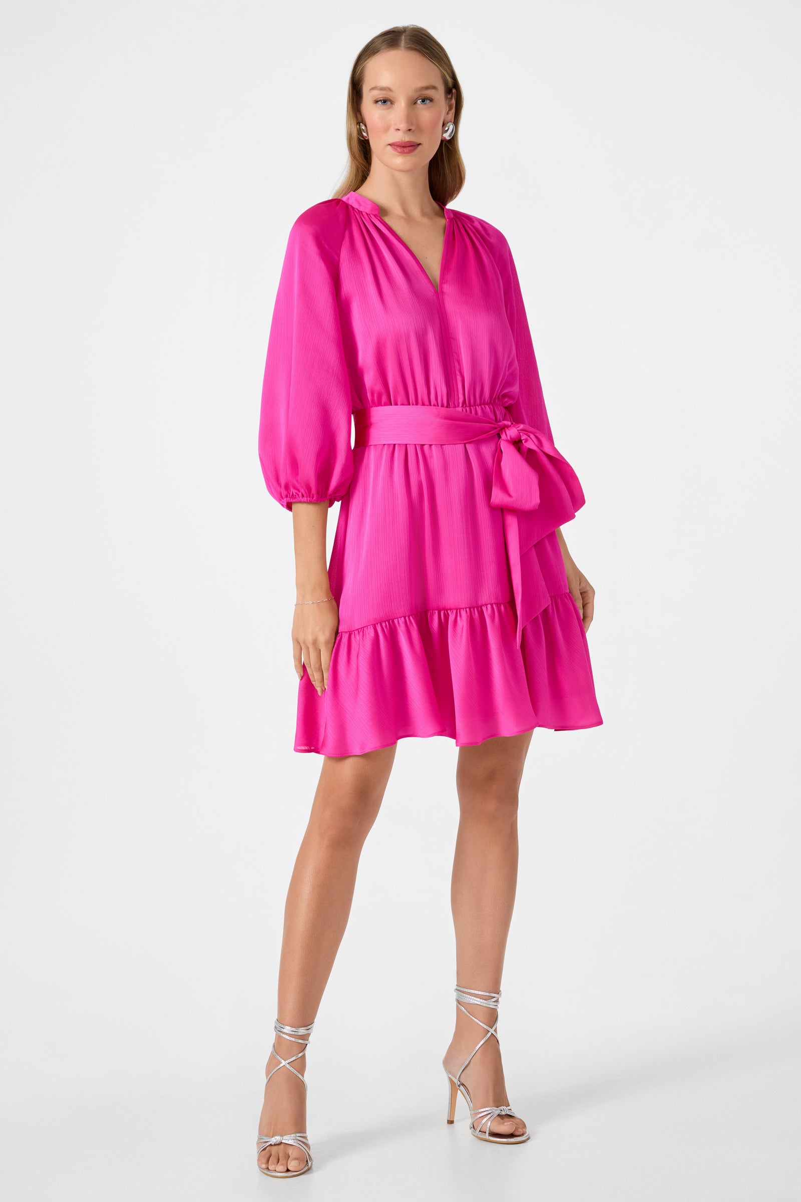 A woman wears the Asuma 3/4 Sleeve Mini Founce Dress in Trina Pink, featuring a v-neckline, waist tie, and ruffled hem. She pairs it with strappy silver high heels and minimal jewelry against a plain white background.