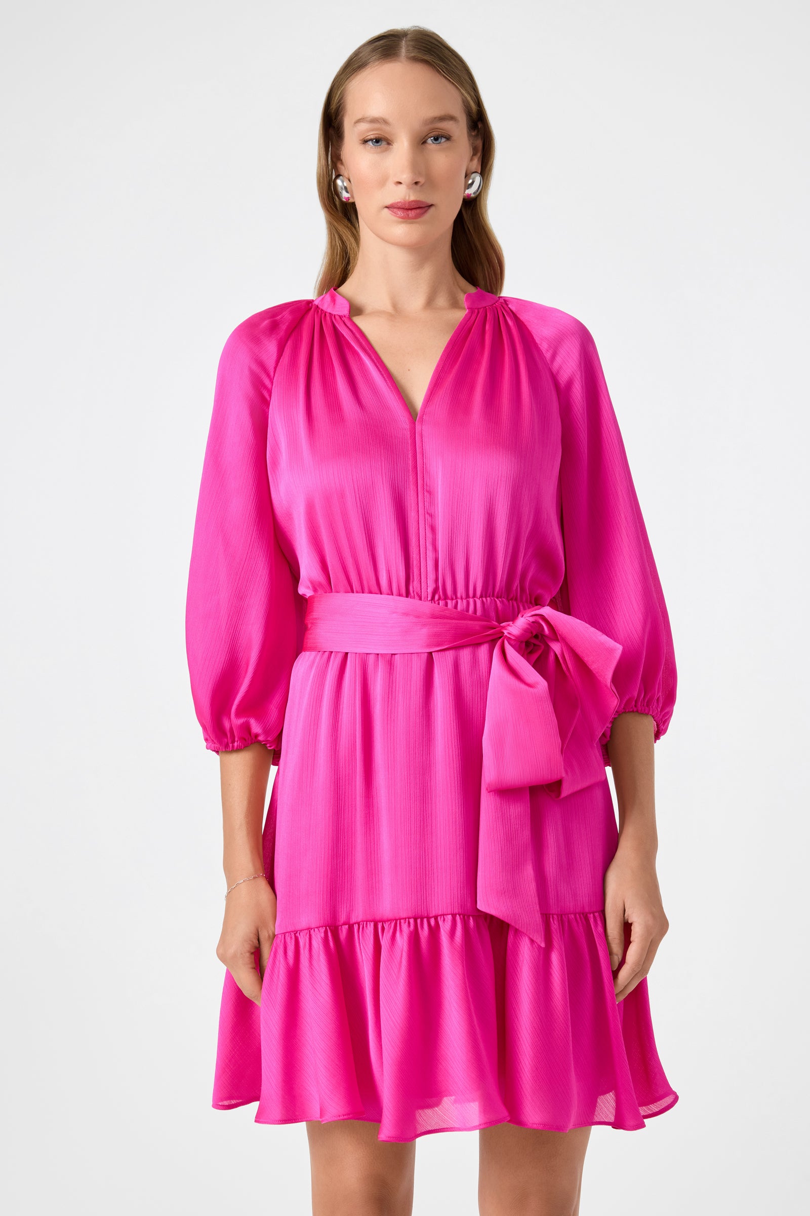 A woman poses against a white background wearing the Asuma 3/4 Sleeve Mini Founce Dress in bright pink, featuring 3/4 sleeves, a cinched waist with bow, and a ruffled hem.