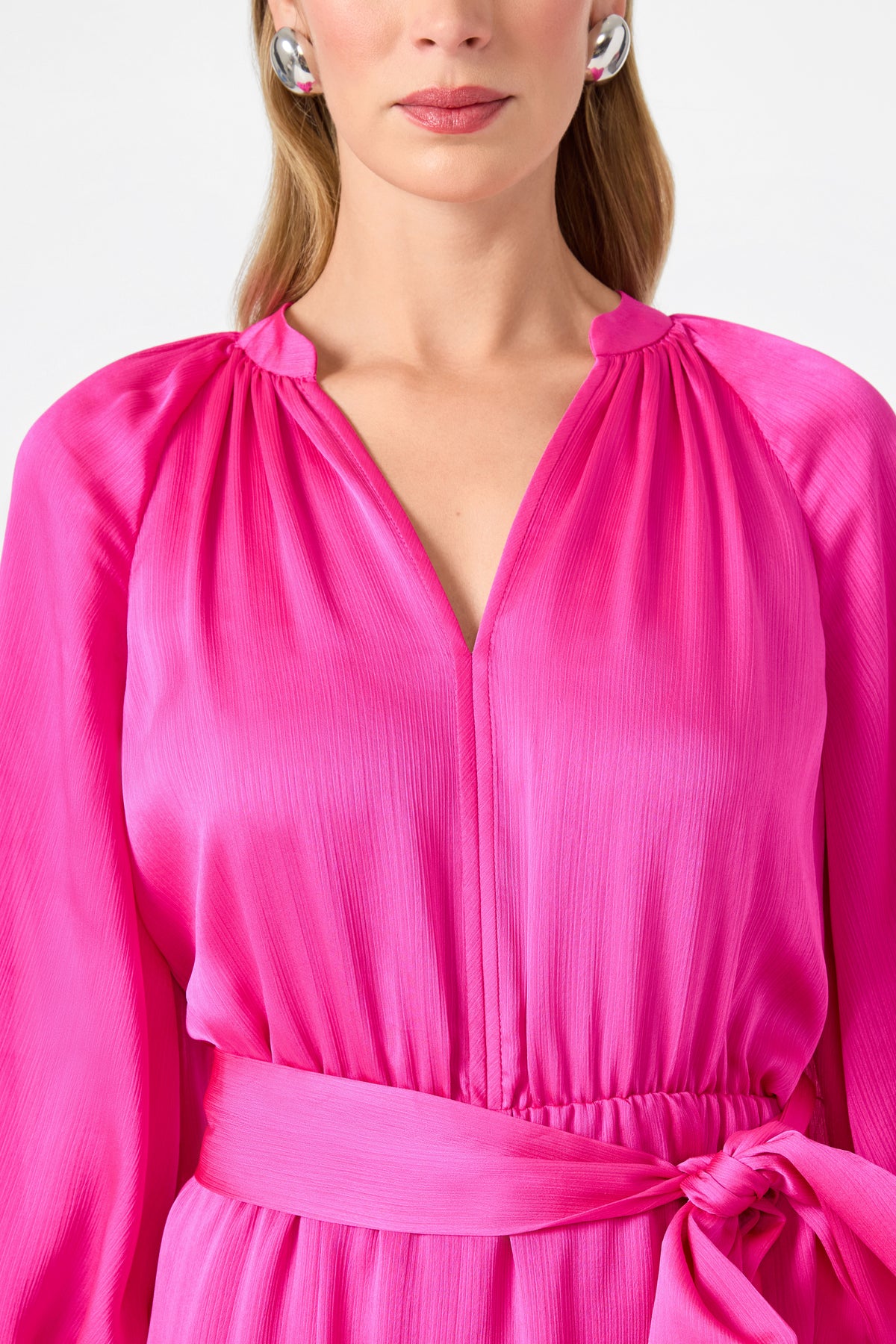 A woman models the Asuma 3/4 Sleeve Mini Founce Dress in vibrant pink, featuring a belted waist, pleated texture, and V-neckline. Silver statement earrings and light pink lipstick complete her look in a cropped photo from shoulders to waist.