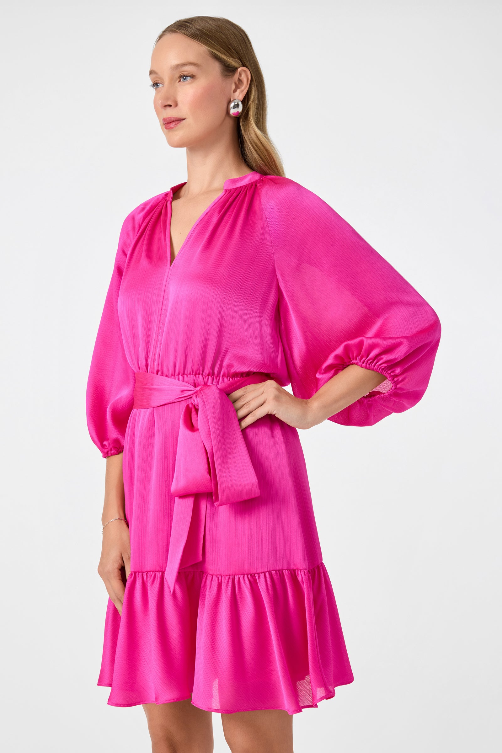 A woman stands against a plain light background wearing the Asuma 3/4 Sleeve Mini Founce Dress in bright pink, featuring puffed sleeves, a V-neckline, waist tie, and ruffle hem.