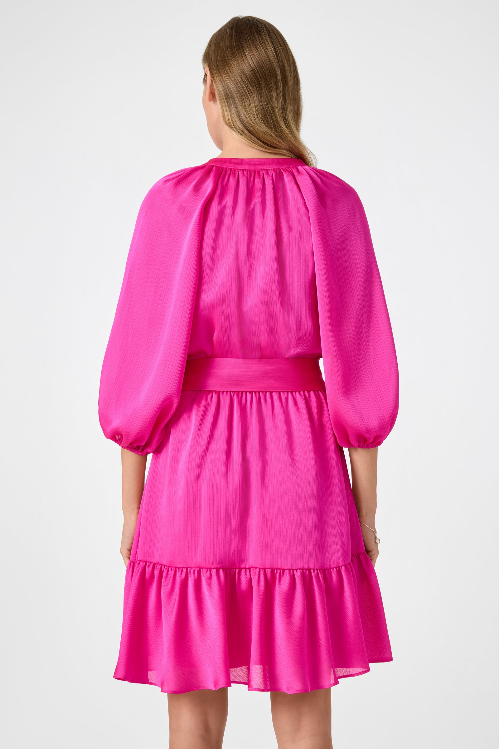 A woman with light brown hair, seen from the back, wears the Asuma 3/4 Sleeve Mini Founce Dress in bright pink with a belted waist and ruffled hem against a plain white background.