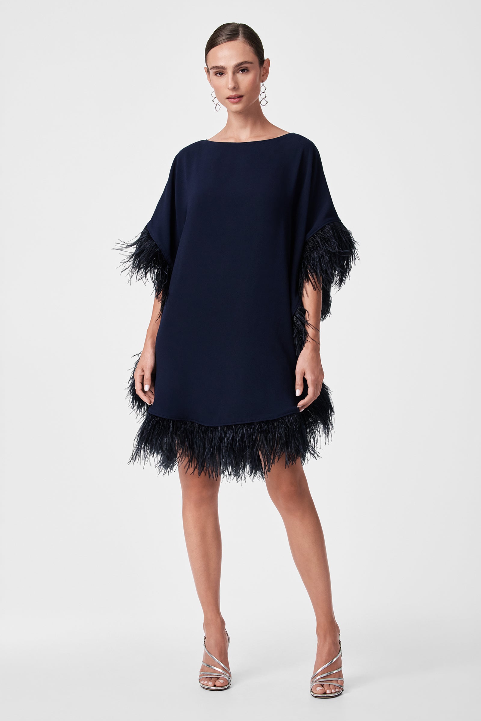 A woman stands against a plain background wearing the Mini Neena Caftan Dress, a loose-fitting navy blue outfit with feathered trim on the sleeves and hem. Her hair is tied back, and she pairs it with silver heels and dangling earrings.