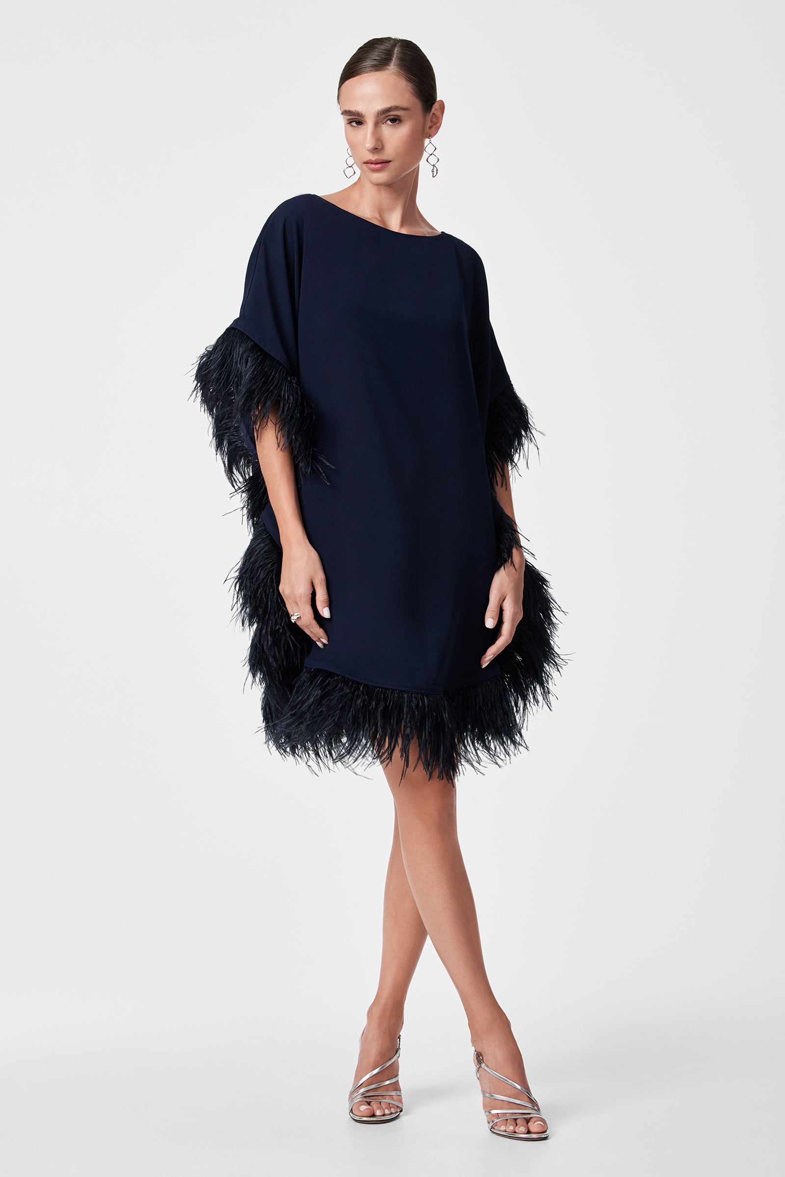 A woman poses against a white background in the Mini Neena Caftan Dress, a navy blue style with feathered trim on the sleeves and hem. She completes the look with silver strappy heels, drop earrings, and neatly pulled-back hair.