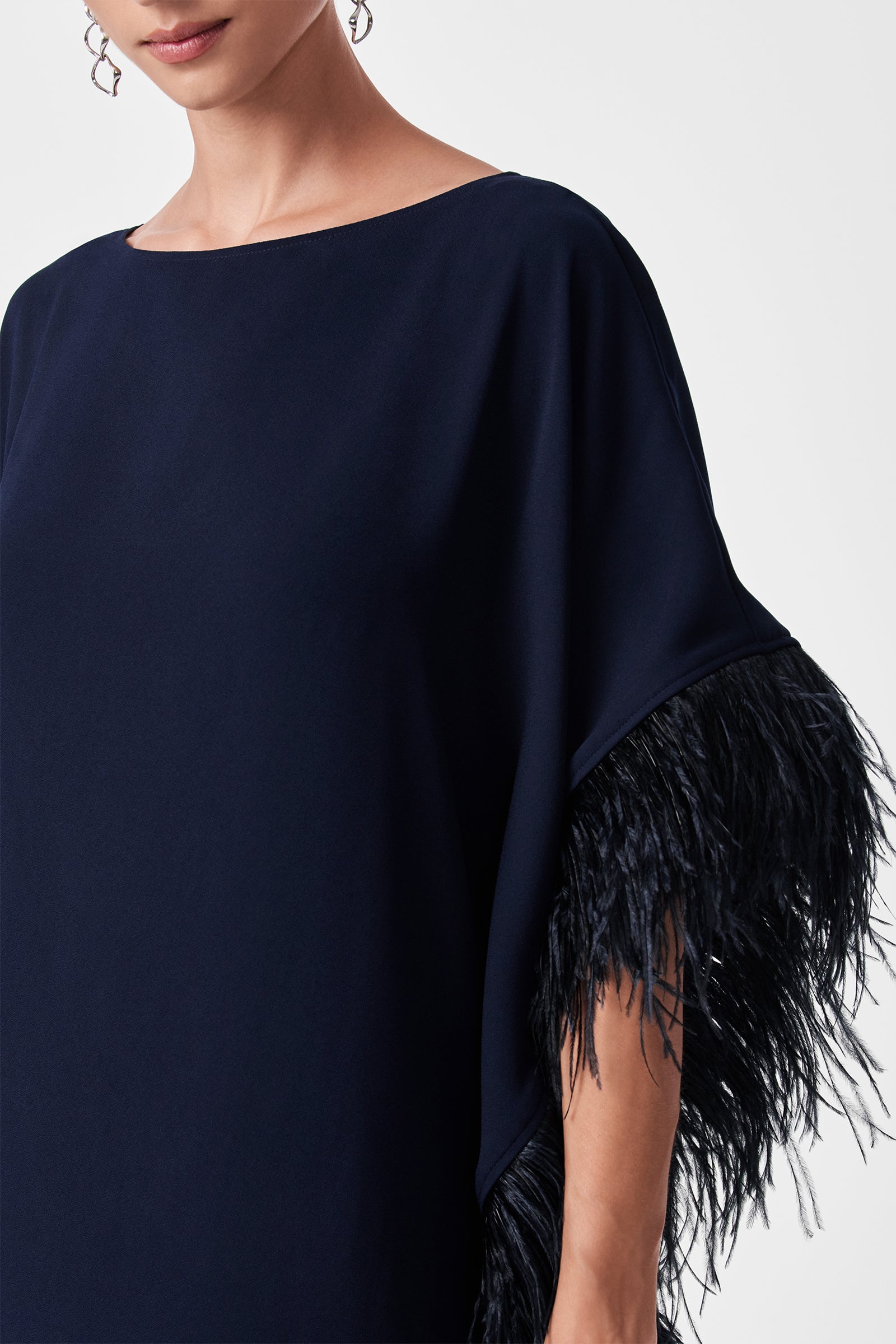 A woman models the Mini Neena Caftan Dress, a navy blue style featuring feathered trim on the sleeve; her lower face, upper body, and one arm appear against a plain background.