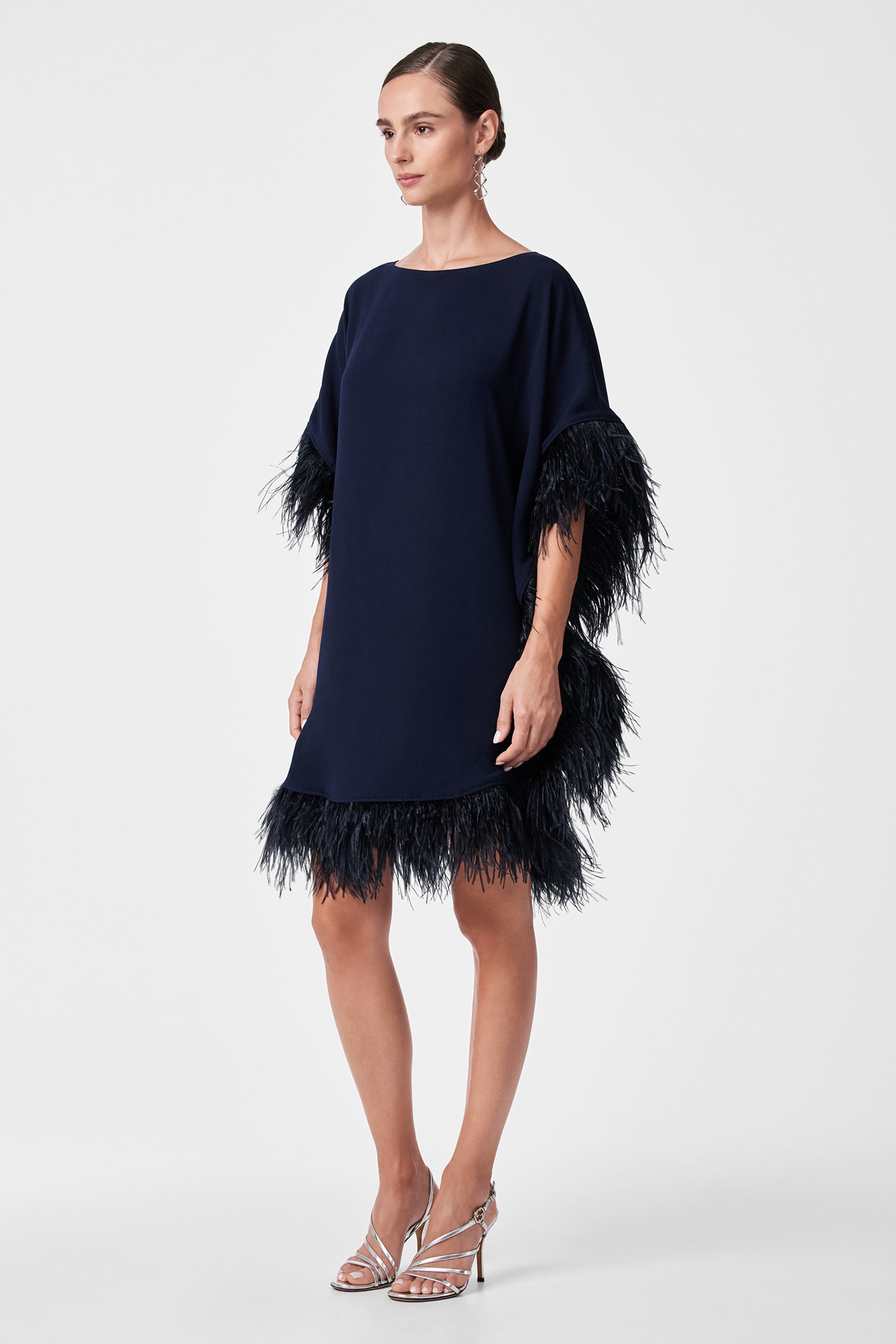 A woman stands against a white background in the Mini Neena Caftan Dress—navy blue, loose-fitting, with feathered trim on the sleeves and hem—paired with silver strappy heels.