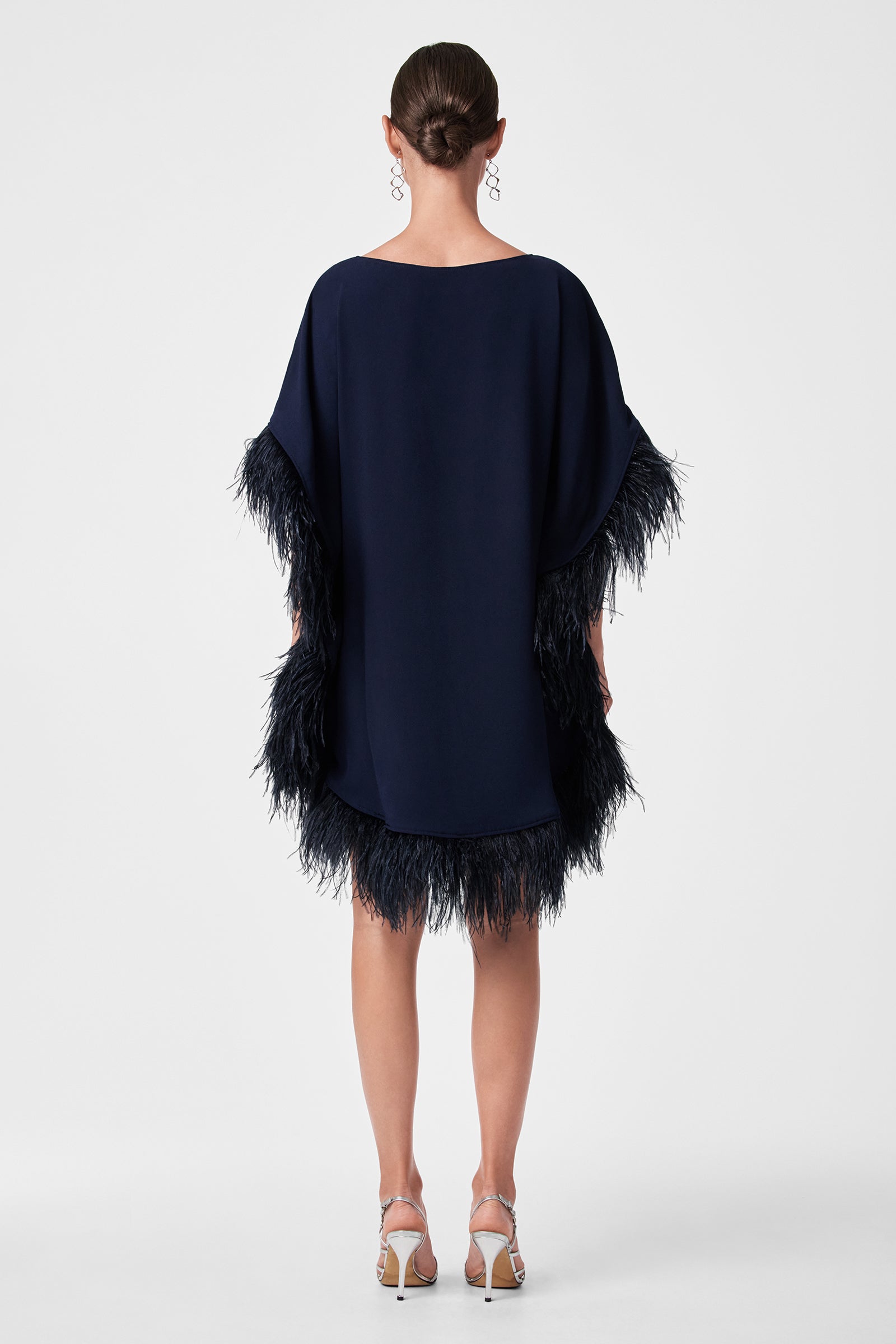 A woman faces away, wearing the Mini Neena Caftan Dress in loose navy blue with feathered trim on sleeves and hem. Her hair is in a neat bun, paired with silver strappy heels and dangling earrings, set against a plain white background.