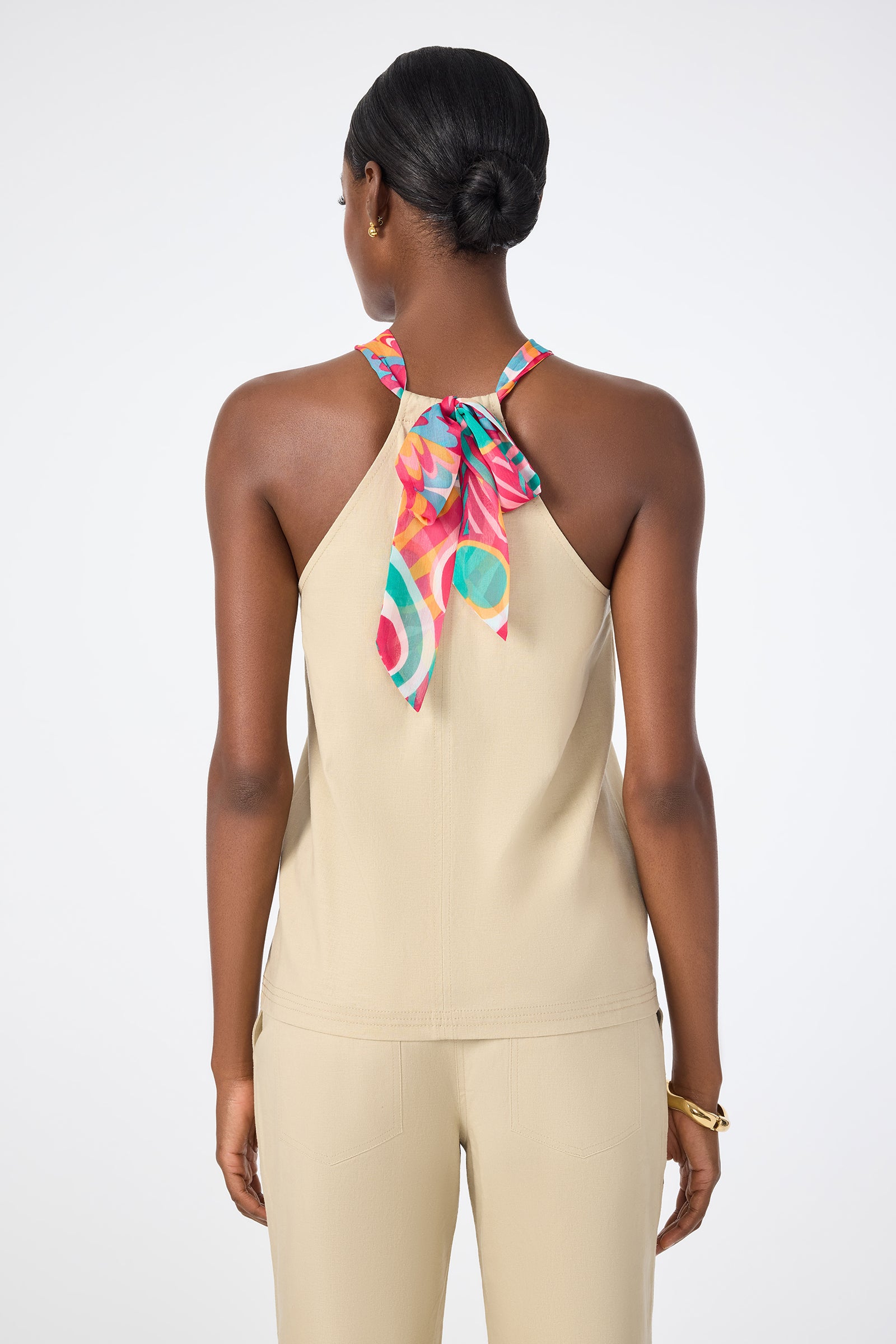A woman faces away, wearing the Chatone Scarf Tie Halter Top in beige with matching pants. The top is tied at the neck with a patterned scarf. Her hair is in a neat bun, and she accessorizes with a gold bracelet.