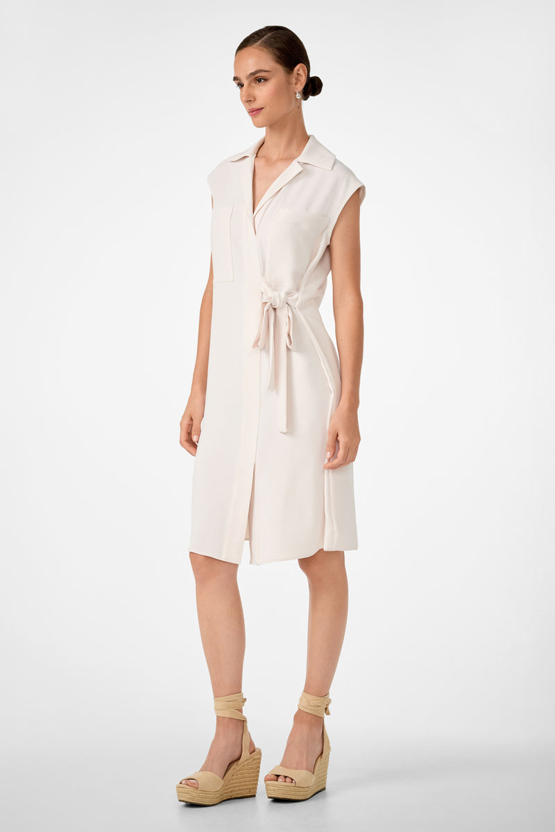 A woman wears the Meron Short-Sleeve Tie-Front Dress, a light beige shirt dress with a waist tie, front pocket, and collar. Her hair is pulled back and she pairs it with beige wedge sandals featuring ankle straps.