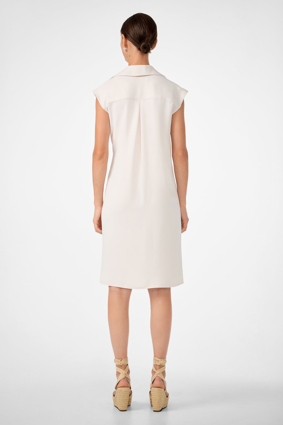 A woman with brown hair in a low bun stands with her back to the camera, wearing the Meron Short-Sleeve Tie-Front Dress and beige wedge sandals with ankle ties, against a plain white background.