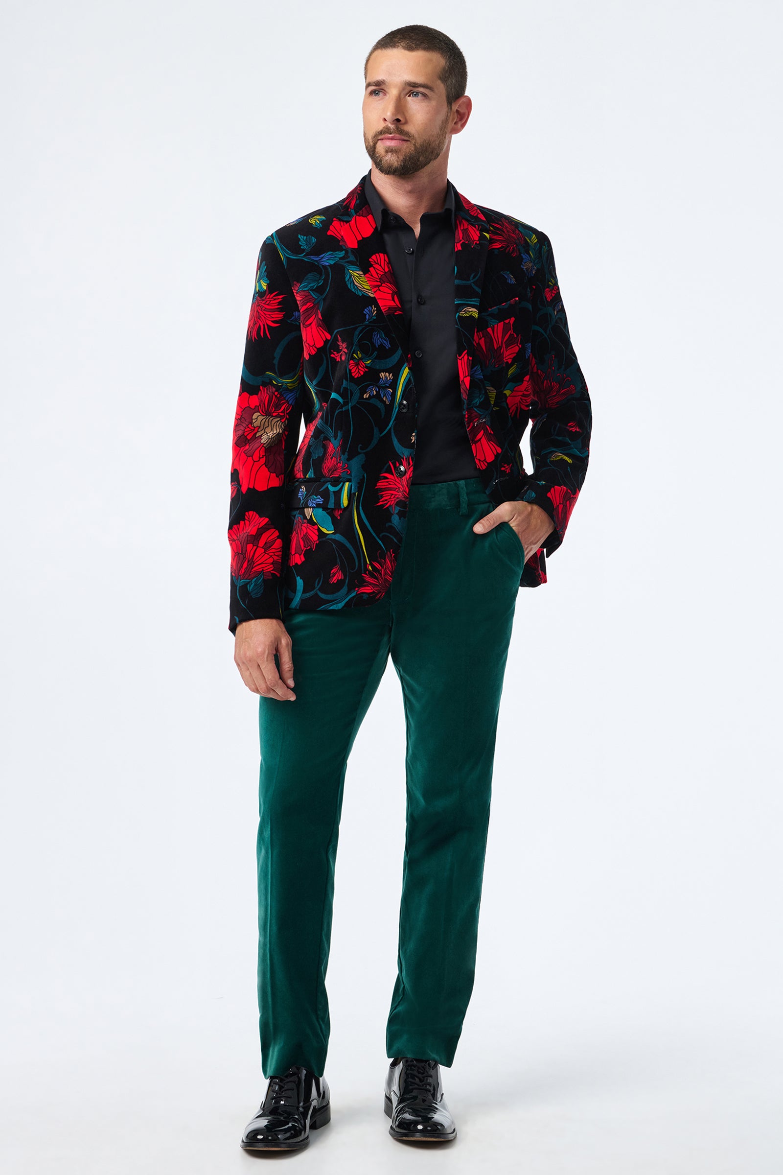 A man stands against a white background wearing black dress shoes, a black shirt, a black blazer with bold red and blue floral patterns, and Clyde Slim Velvet Trouser in green. His left hand is in his pocket.