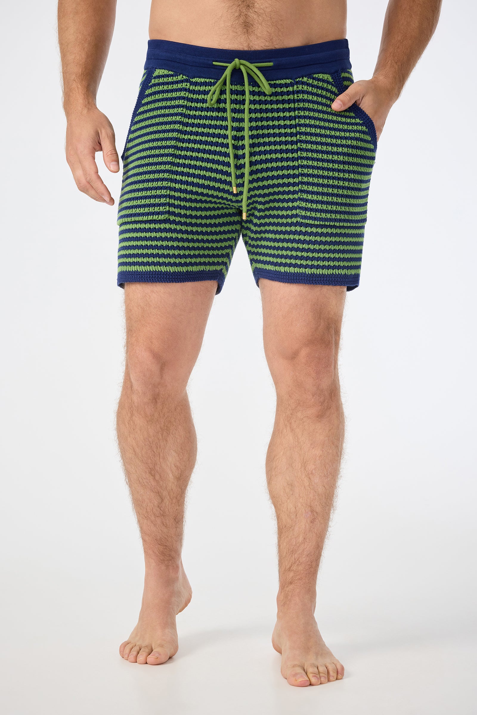 A man stands barefoot with relaxed hands, wearing the August Knit Short in Multi, featuring a blue and green pattern, drawstring waist, and pockets. The image is cropped at his shoulders.