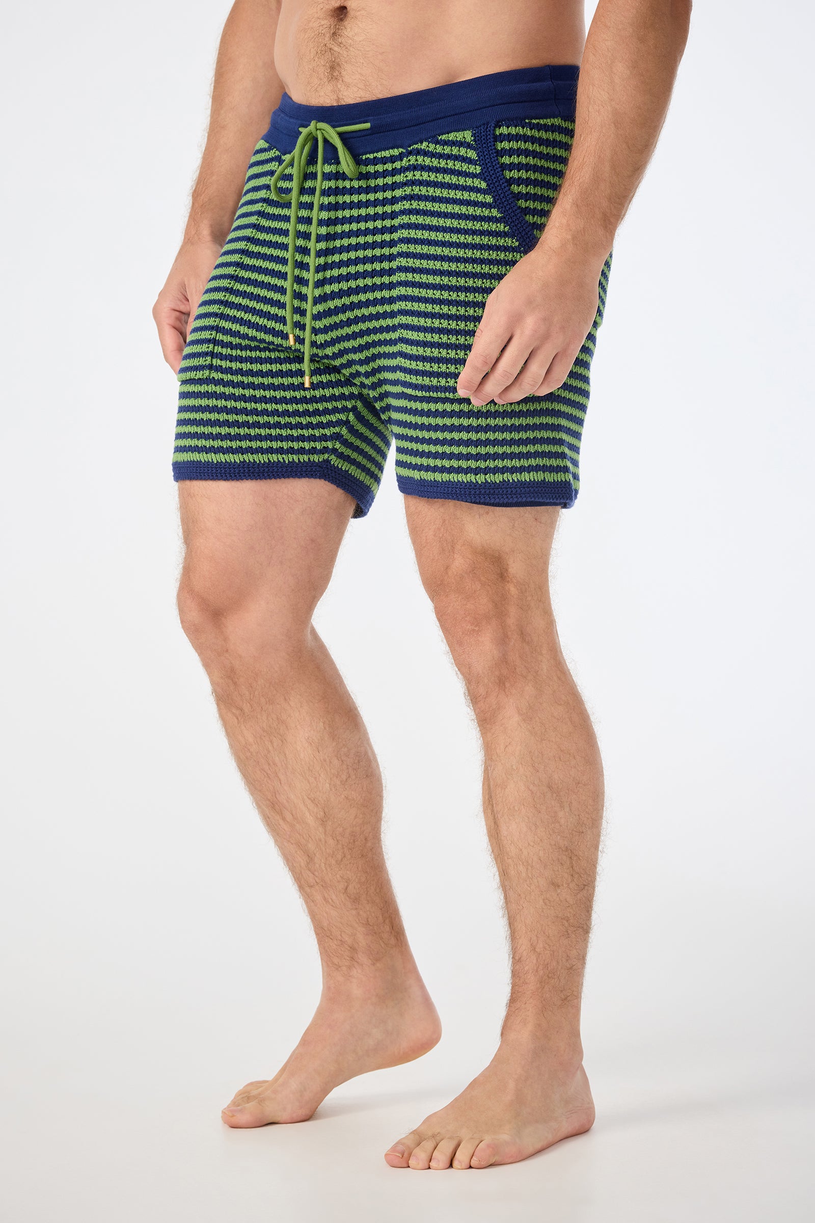 A man stands barefoot against a plain white background, wearing the August Knit Short featuring a green and blue pattern, a blue waistband, and a green drawstring.