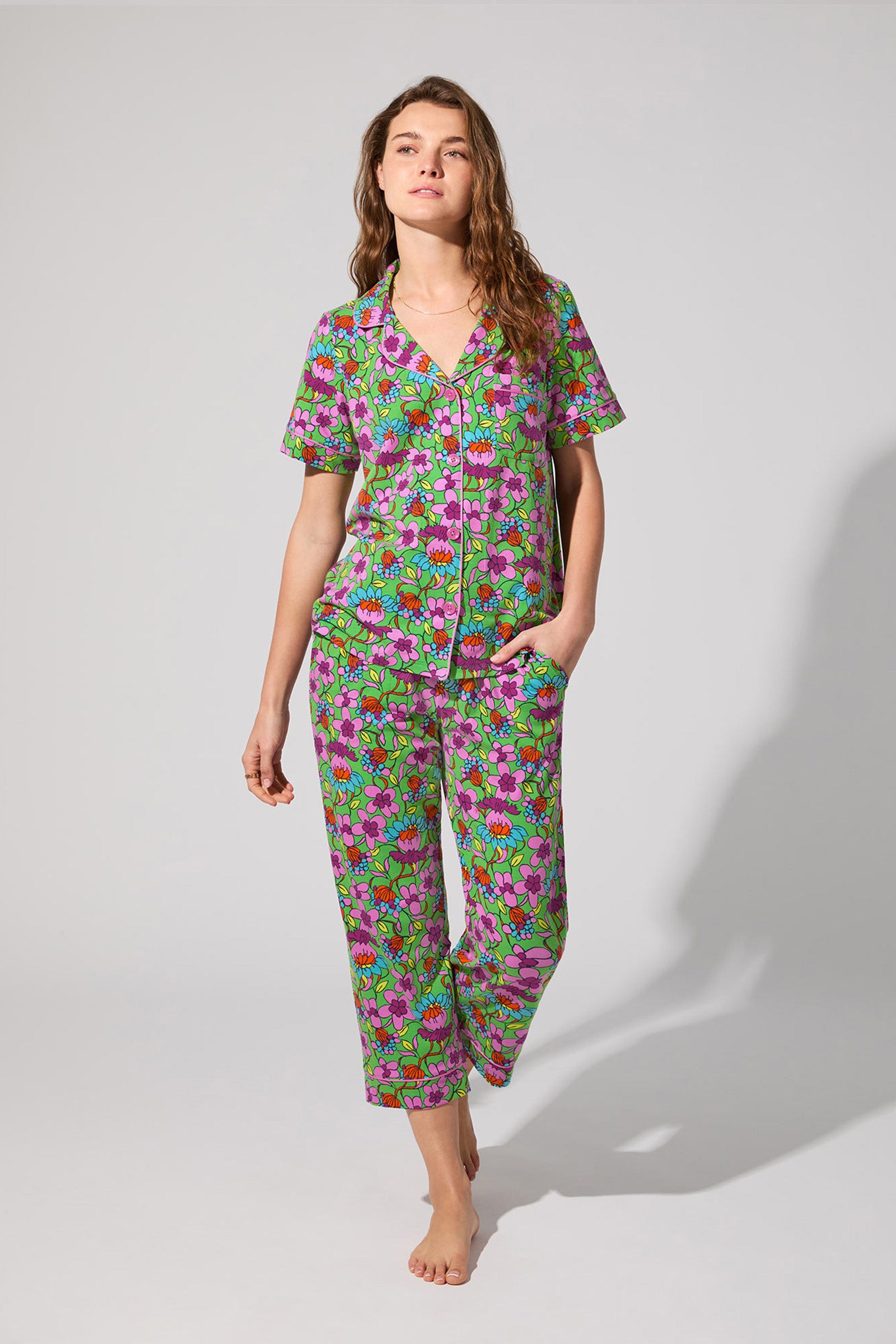 A woman stands barefoot against a plain background, wearing the Crop Classic PJ Set in Garden Floral. The short-sleeved, button-up set features a colorful green, pink, and purple floral pattern. One hand is in her pocket as she looks to the side.
