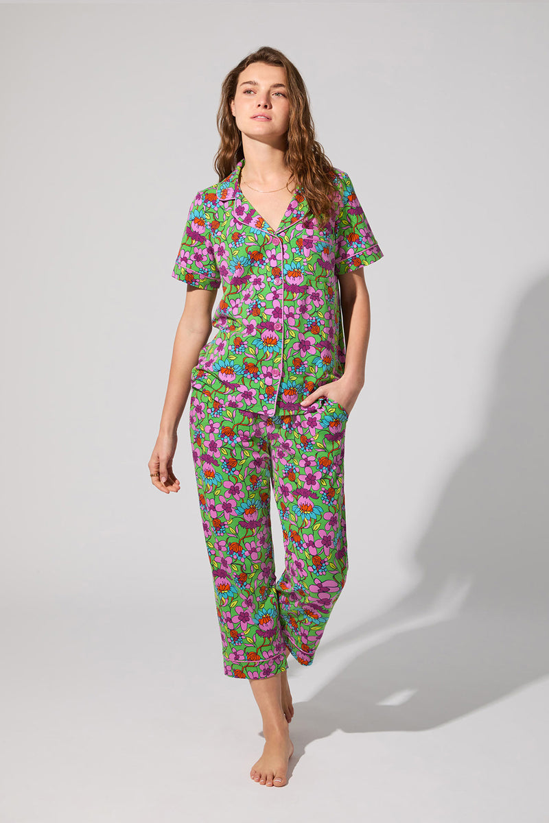 A woman stands barefoot against a plain background, wearing the Crop Classic PJ Set in Garden Floral. The short-sleeved, button-up set features a colorful green, pink, and purple floral pattern. One hand is in her pocket as she looks to the side.