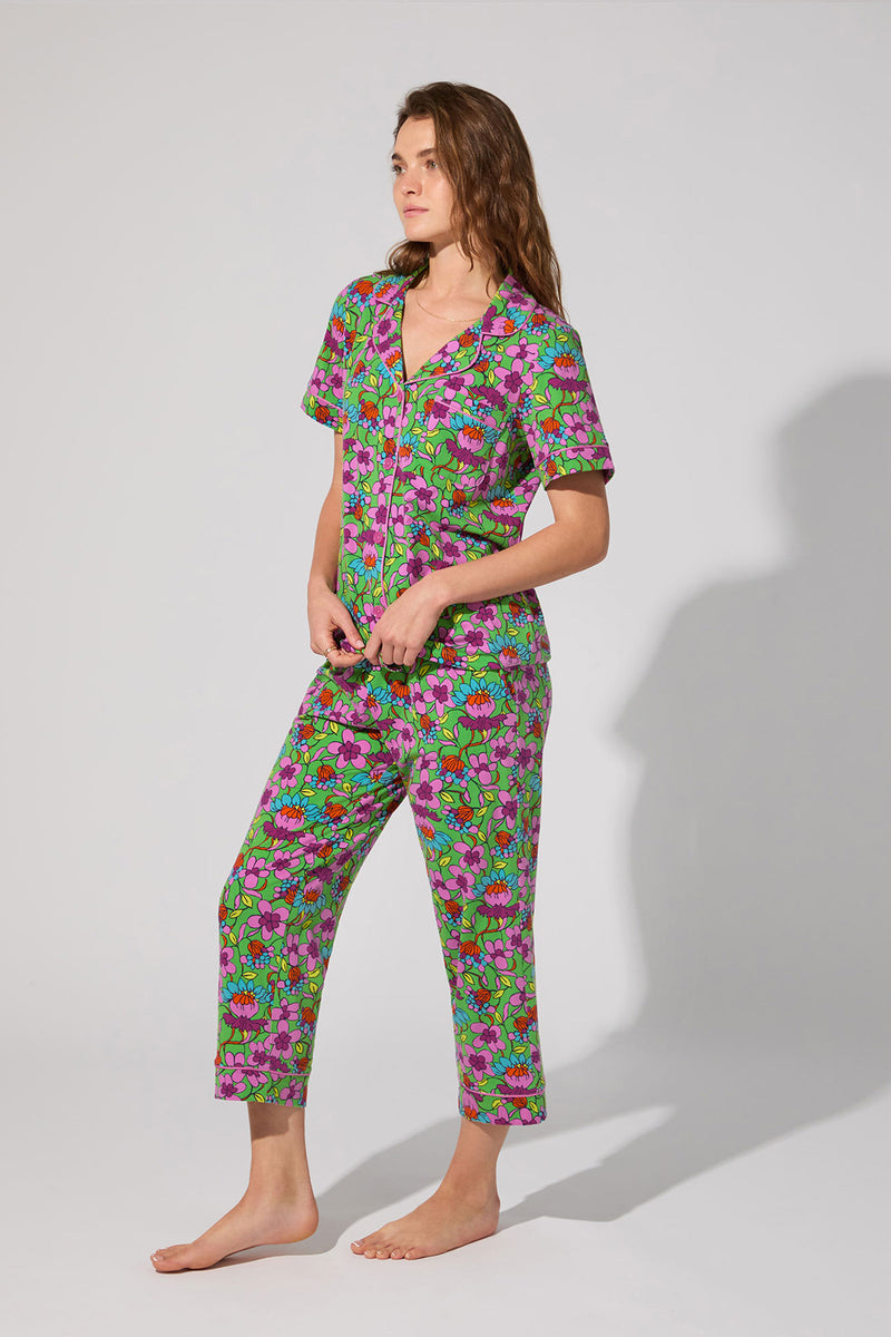 A woman stands barefoot against a plain light background, wearing the Crop Classic PJ Set - Garden Floral, featuring colorful floral-patterned short sleeves and cropped pants. She looks to the side with a relaxed posture.