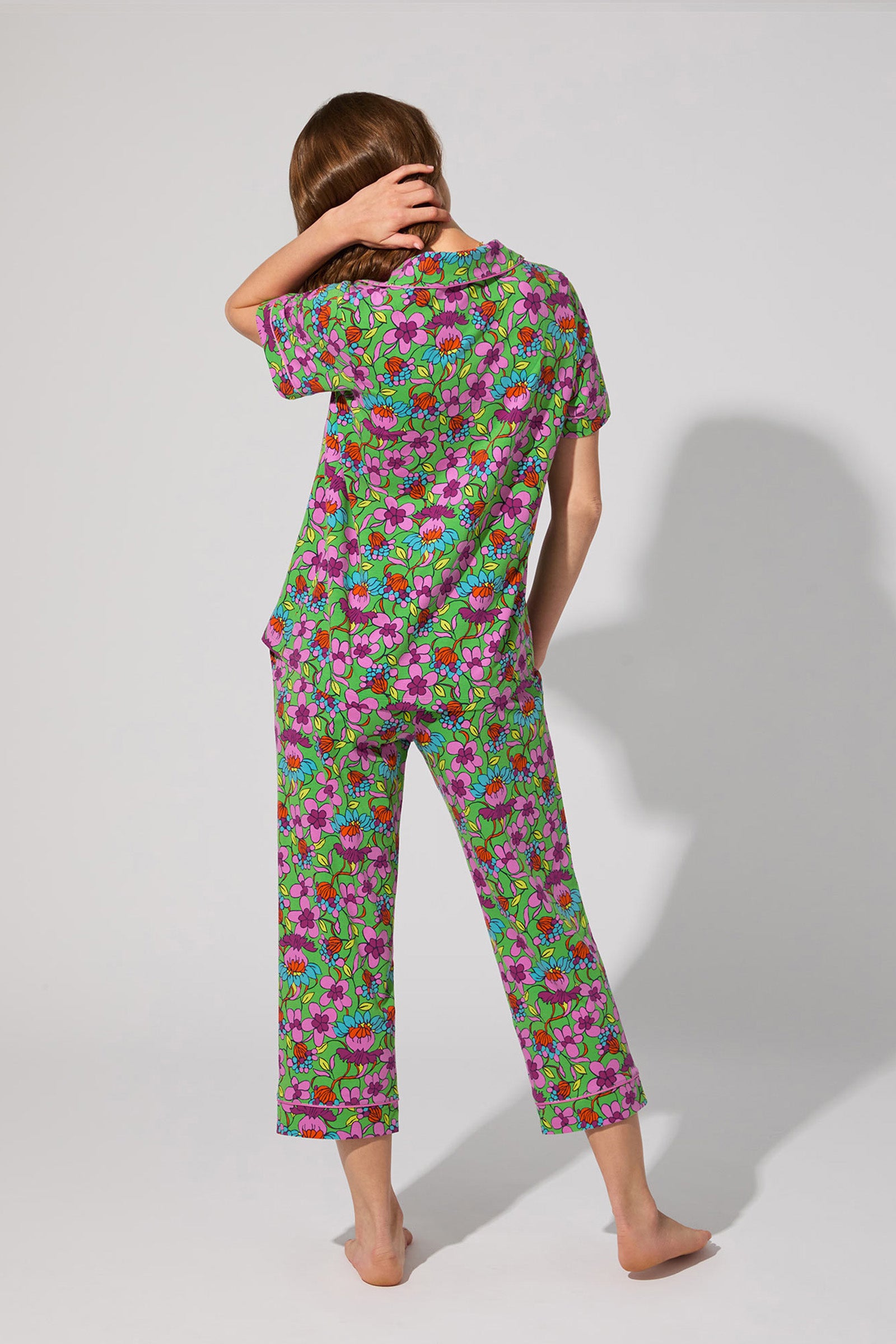 A person stands barefoot, back to camera, wearing the Crop Classic PJ Set - Garden Floral with short sleeves and cropped pants, touching their head with one hand.