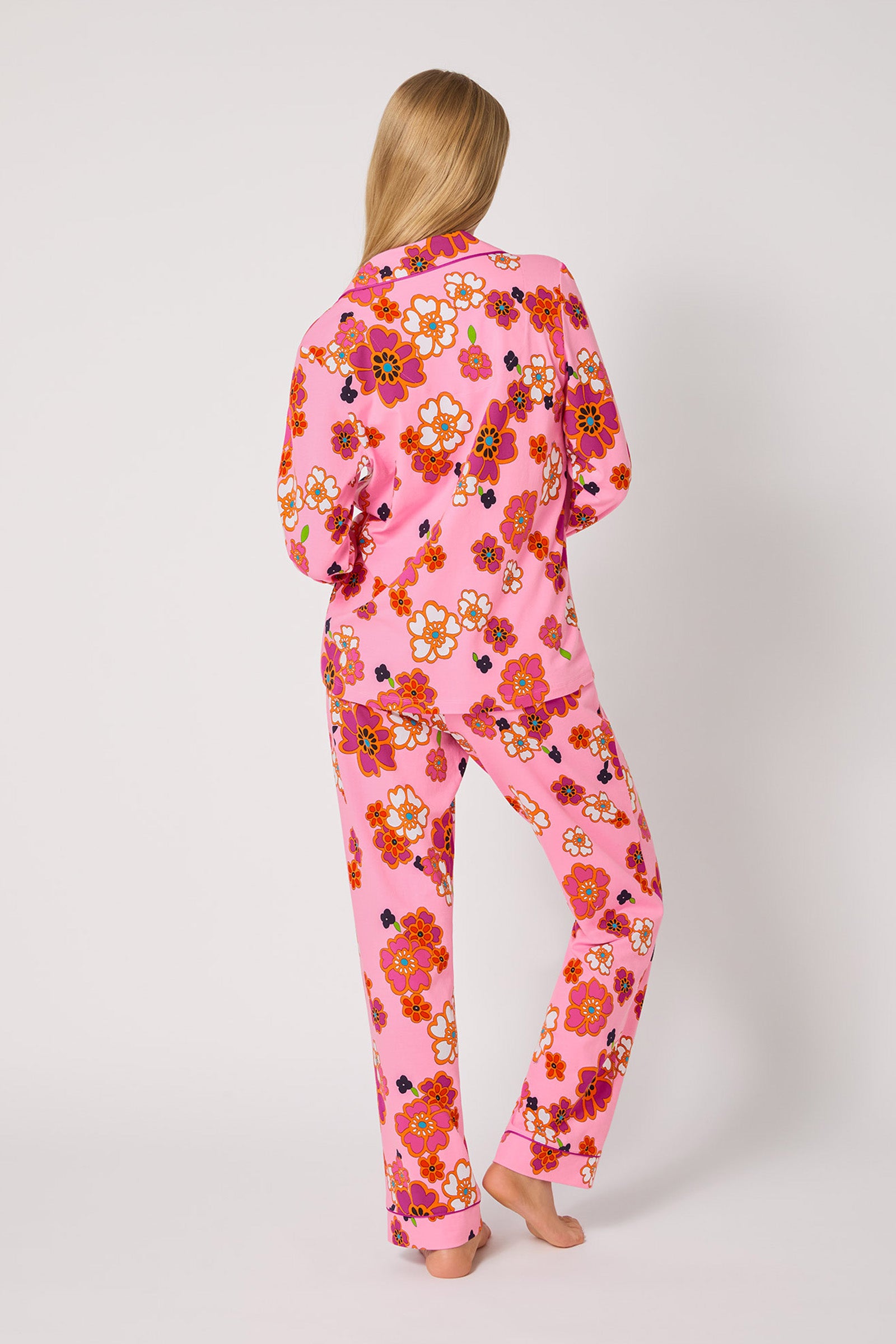 A person with long blonde hair stands barefoot, facing away, wearing the Resort Floral L/S Classic Pj—a pink pajama set featuring a vibrant floral pattern. The background is plain white.