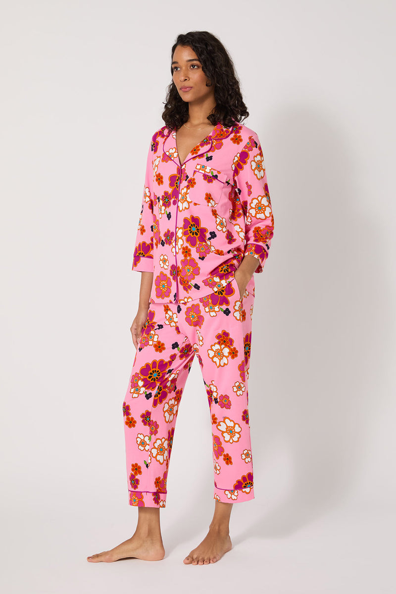 A woman stands barefoot in Resort Floral 3/4 Cropped Pj, featuring pink, red, orange, and white floral print. The pajama set has long sleeves, cropped pants, a button-up front, and is shown against a plain white background.