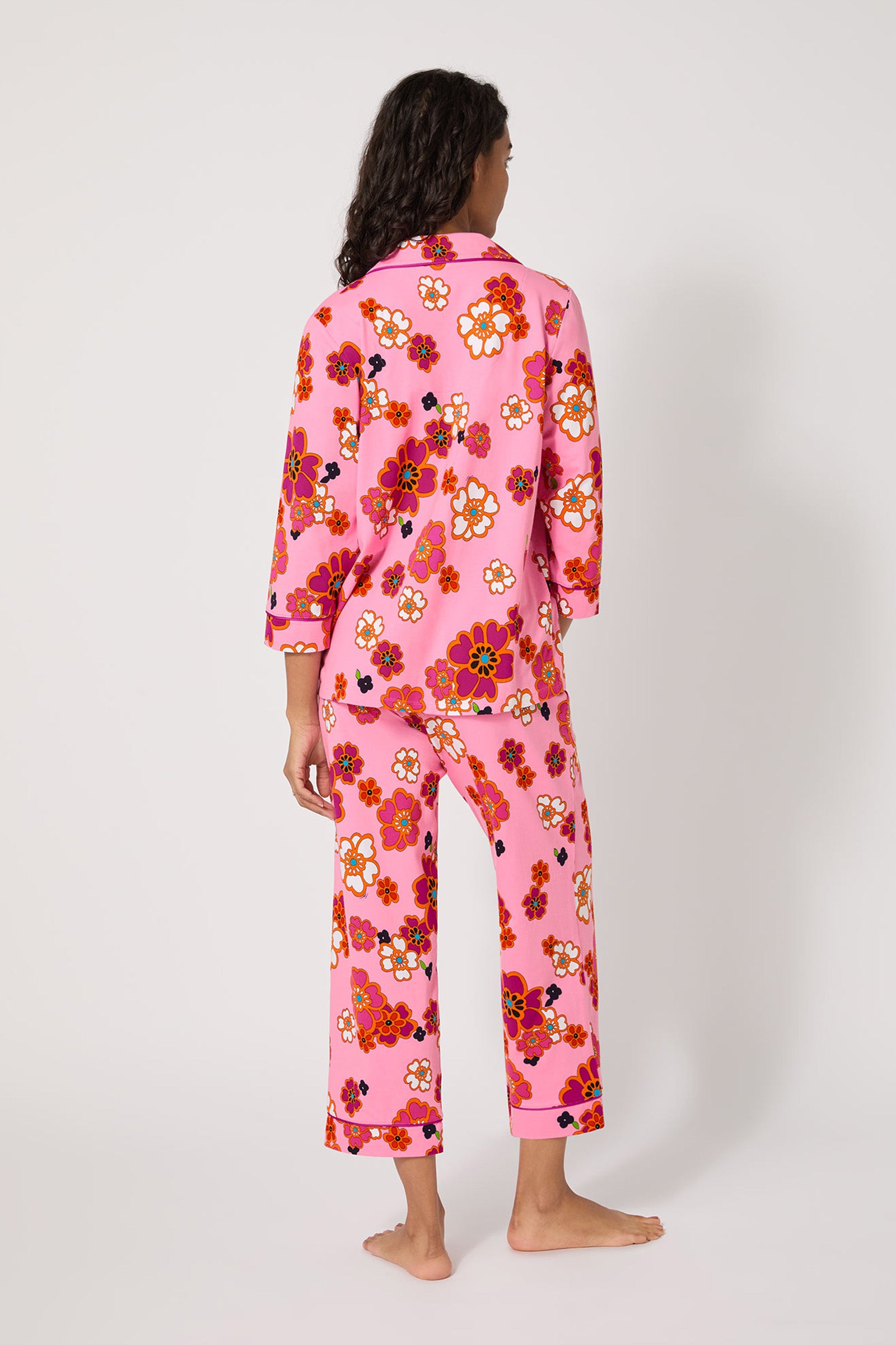 A person with long dark hair stands barefoot facing away, wearing the Resort Floral 3/4 Cropped Pj—pink pajamas featuring a bold multicolored floral print—against a plain white background.