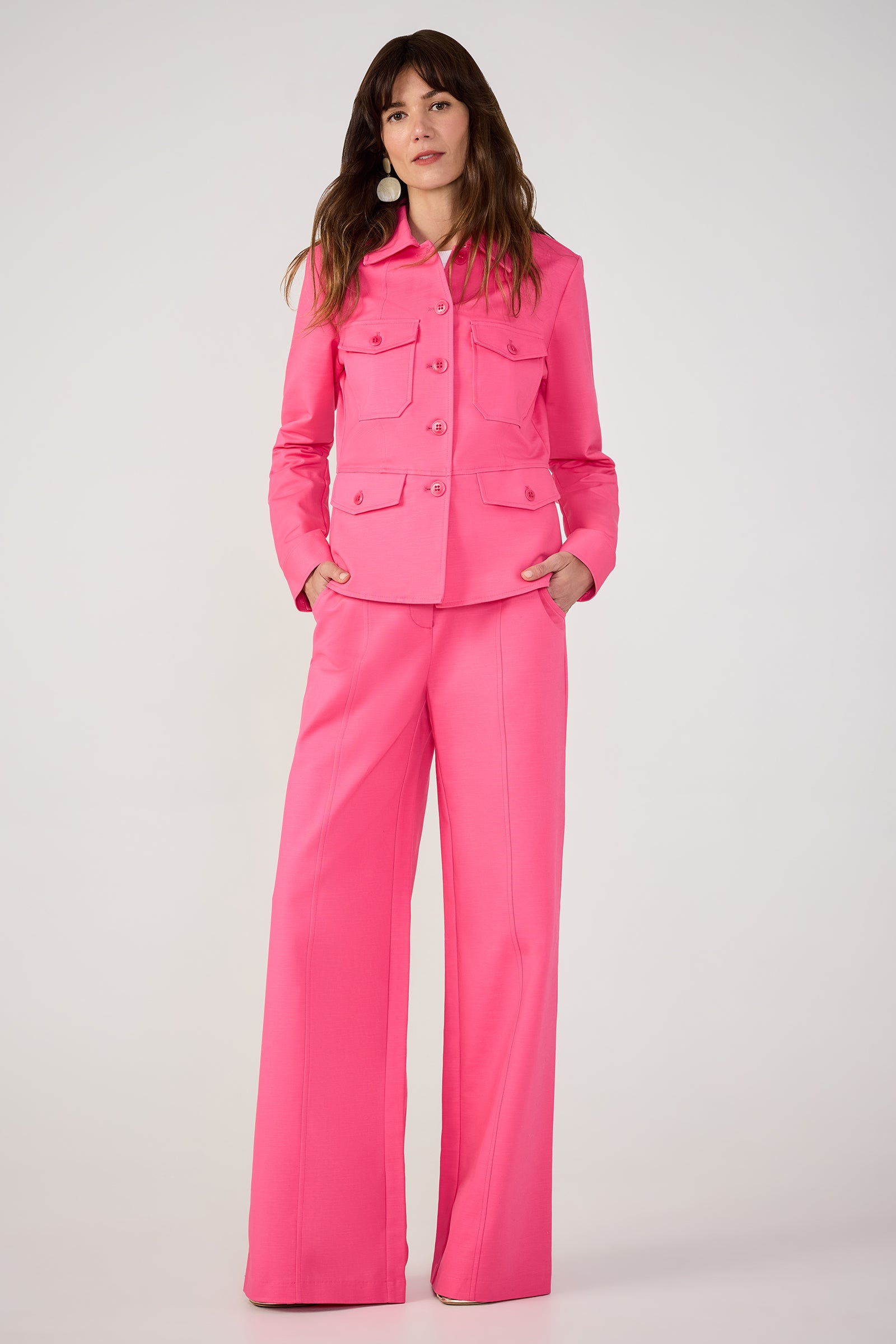 Woman with shoulder-length brown hair stands against a plain background, wearing the Qualifier Button-Front Peplum Jacket - Solid Suiting in bright pink with matching wide-leg trousers, hands in her pockets.