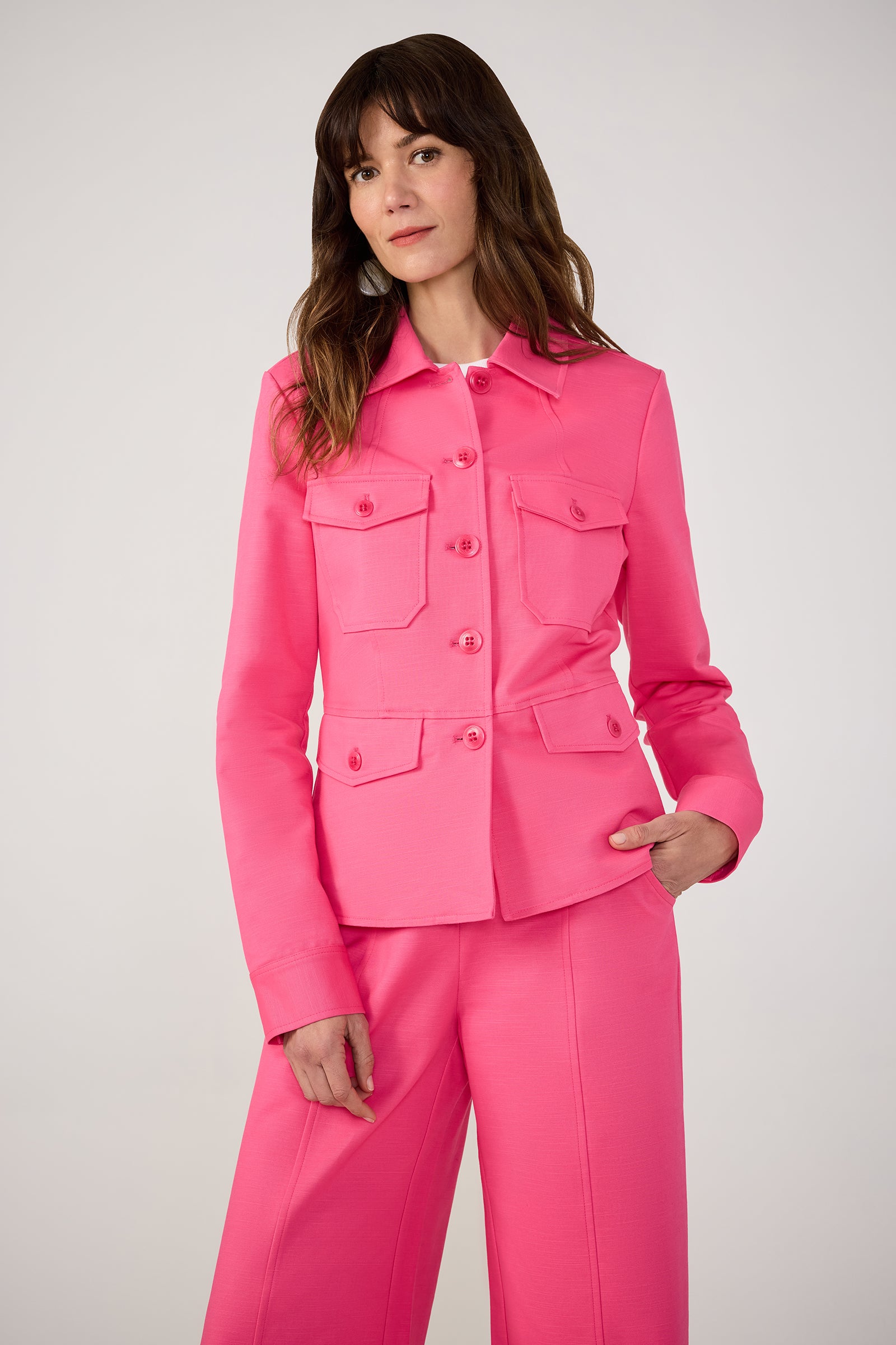 A woman with long brown hair models the Qualifier Button-Front Peplum Jacket - Solid Suiting in Love All Pink, paired with matching pink pants, against a simple light background.