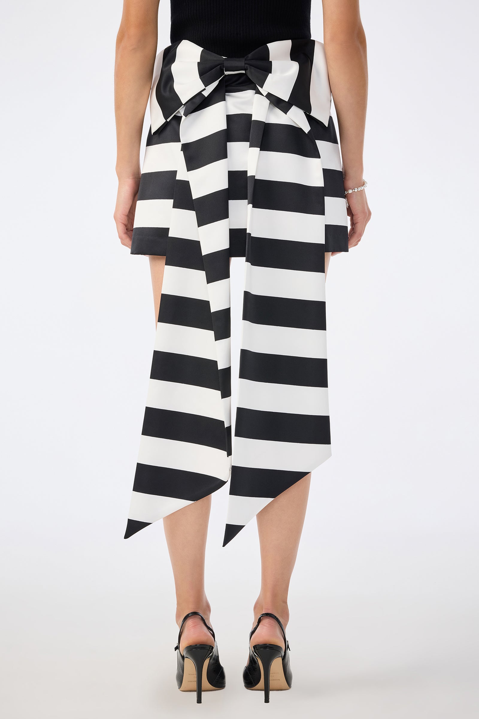 A person stands facing away, wearing a black top and the Vinna Skirt—a black-and-white striped skirt with an oversized bow and long pointed tails at the back, paired with black heels and a bracelet.