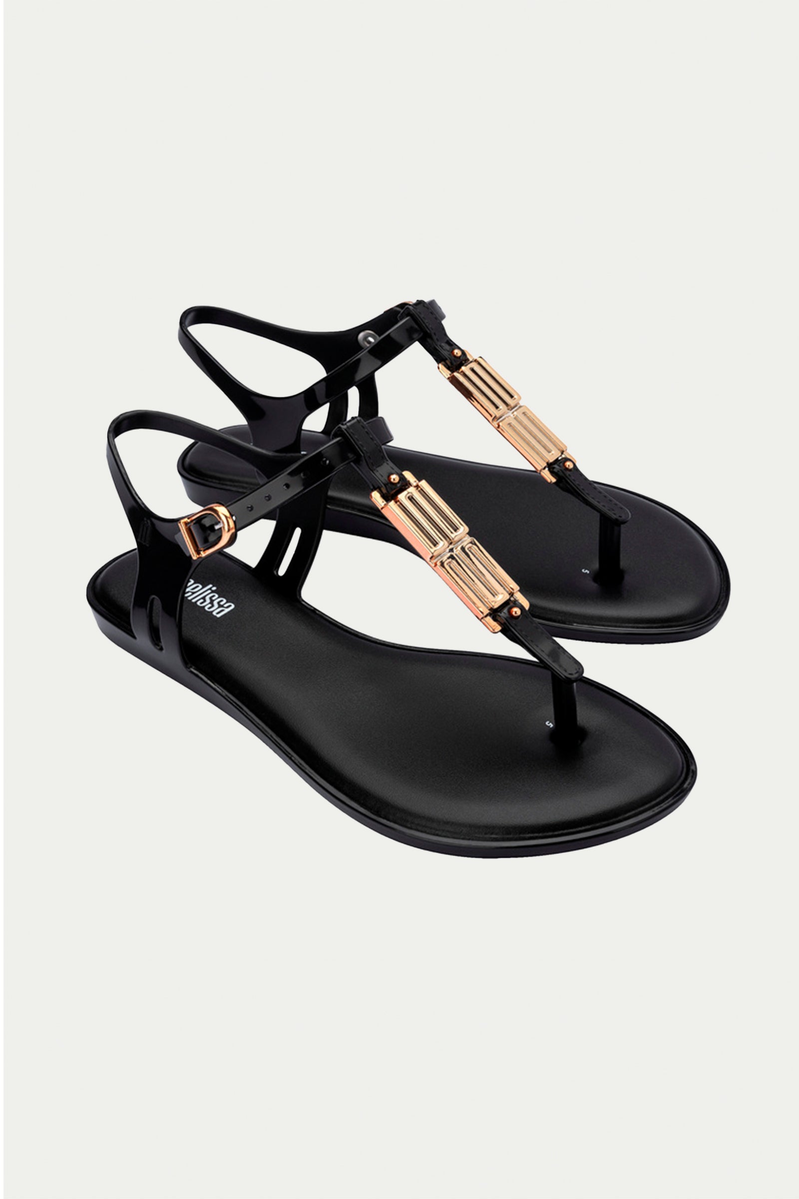 The Melissa Solar Chic T-Strap Sandal in black features gold decorative accents and adjustable ankle straps, elegantly displayed on a plain light background.