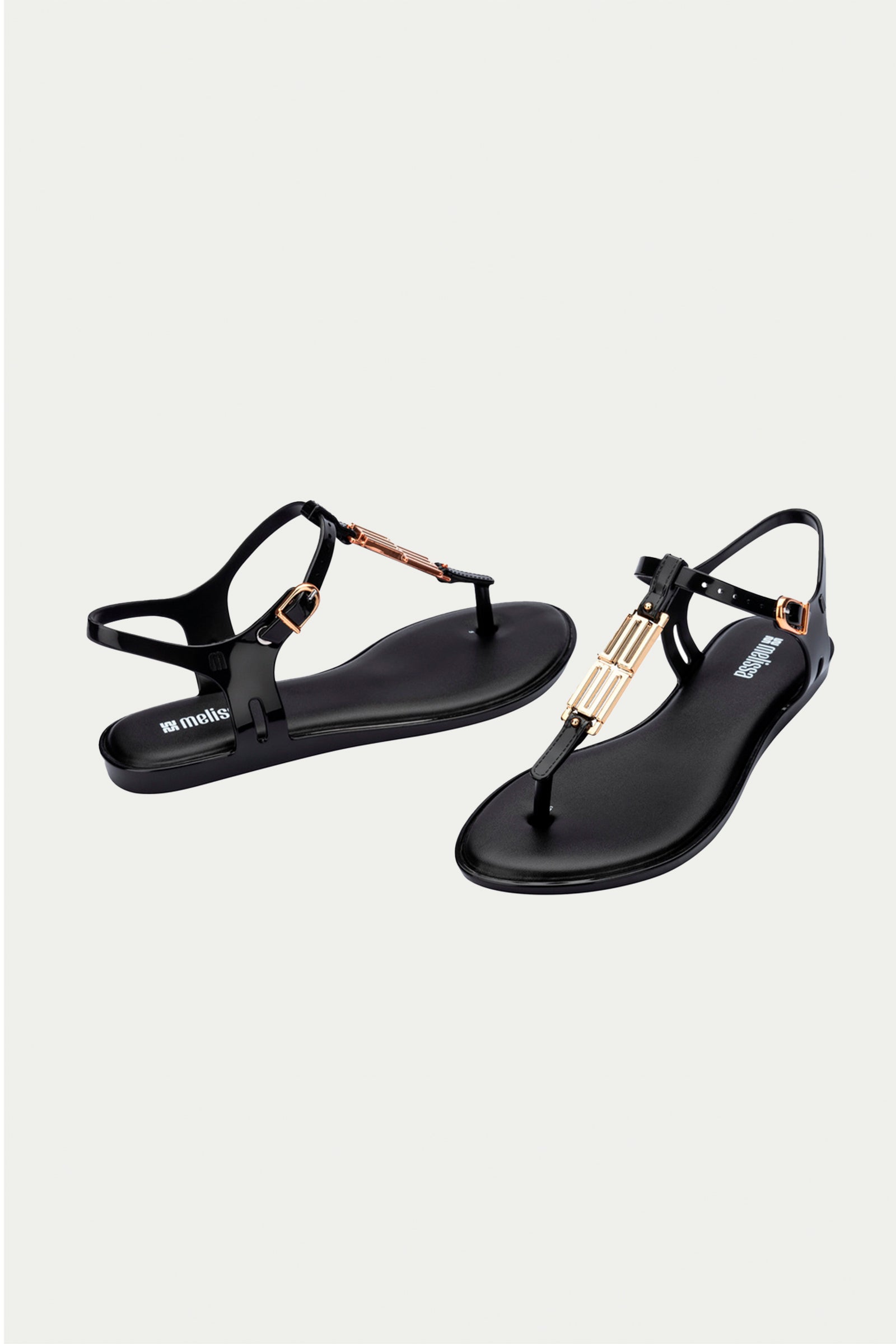 The Melissa Solar Chic T-Strap Sandal in black features thong straps with gold buckle and bar accents, shown against a plain white background.
