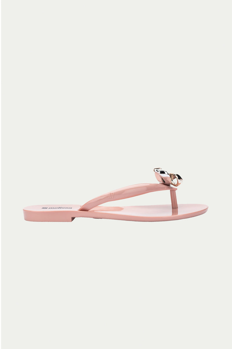 The Melissa Harmonic Eclipse Bow Sandal in pink features a light pink thong strap design with a metallic bow, shown in profile on a plain white background.