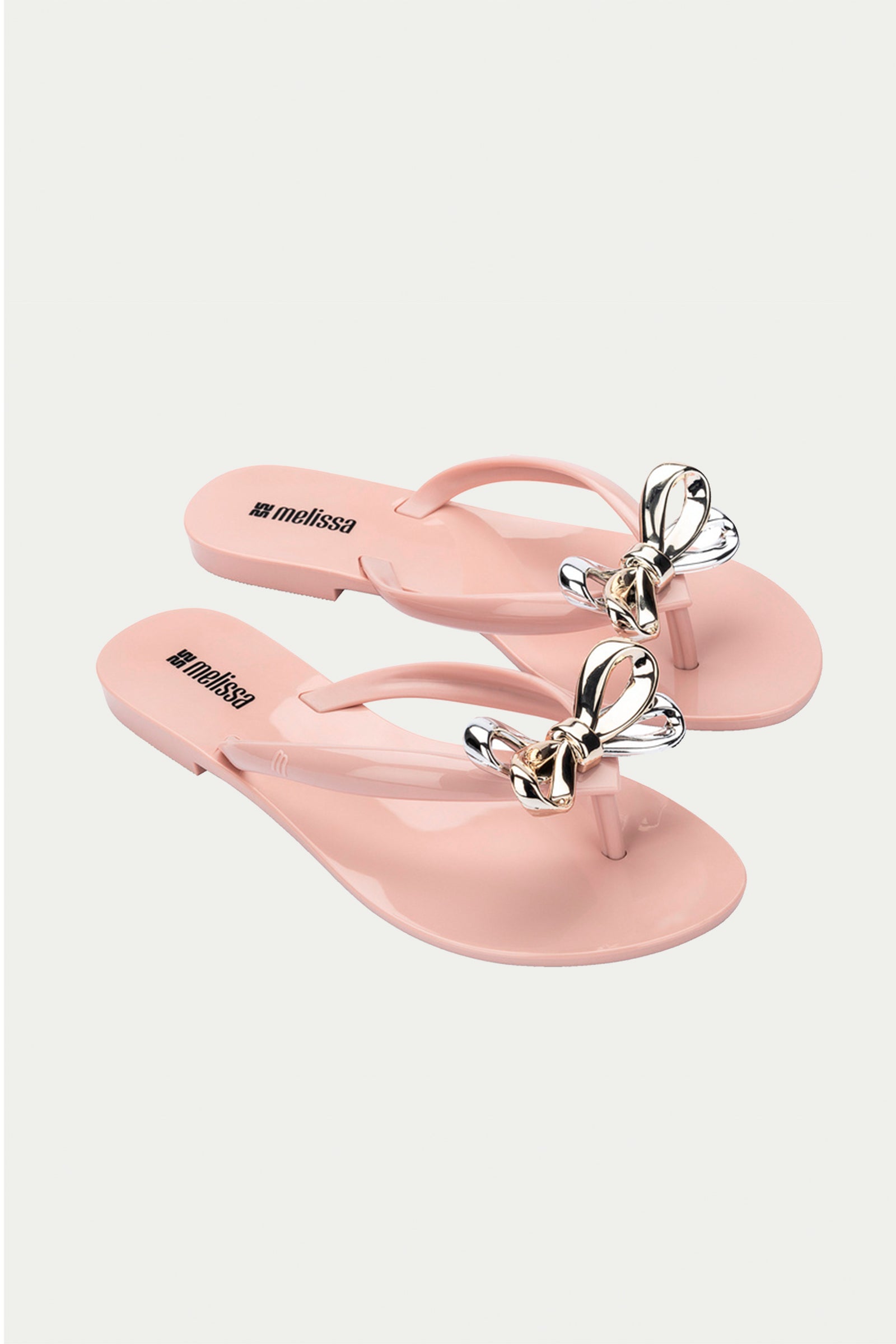 The Melissa Harmonic Eclipse Bow Sandal in pink is a pair of light pink thong sandals featuring metallic butterfly accents on the straps, shown against a white background.