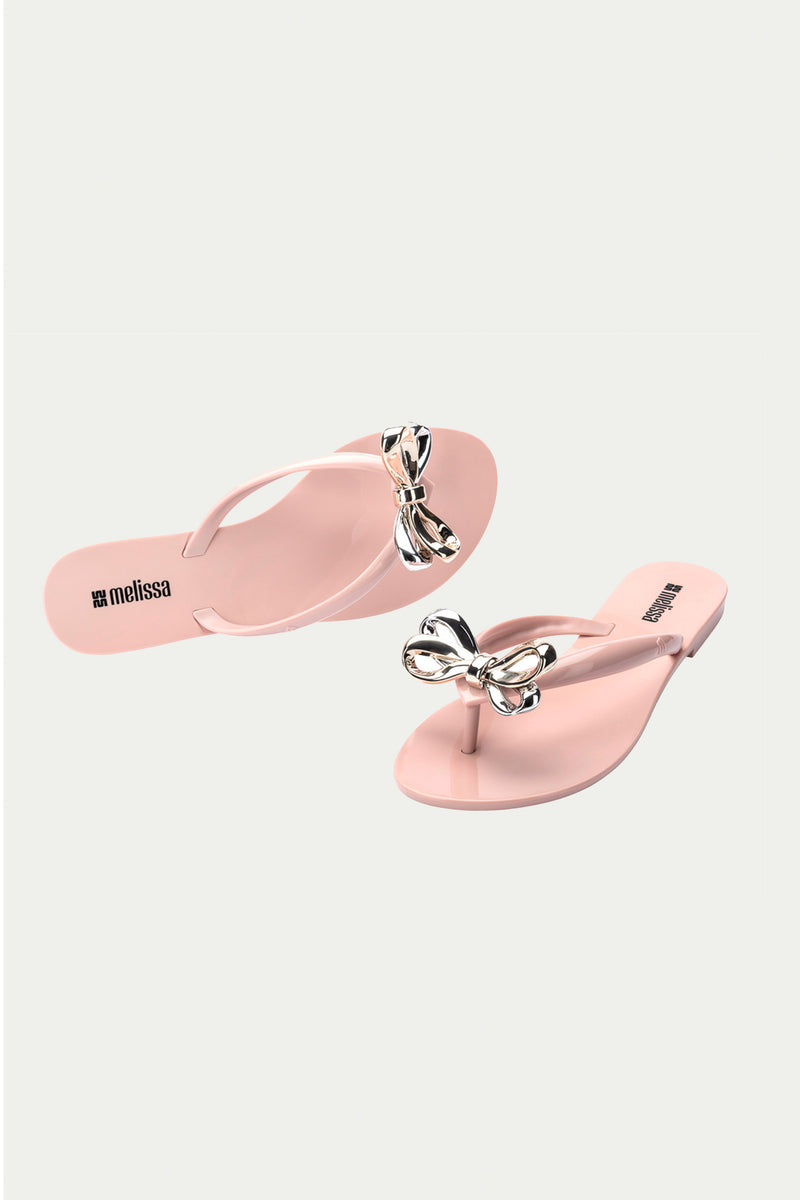 Two light pink Melissa Harmonic Eclipse Bow Sandals with metallic silver bows on the straps, shown on a white background. Melissa is printed in black on each sandals footbed.