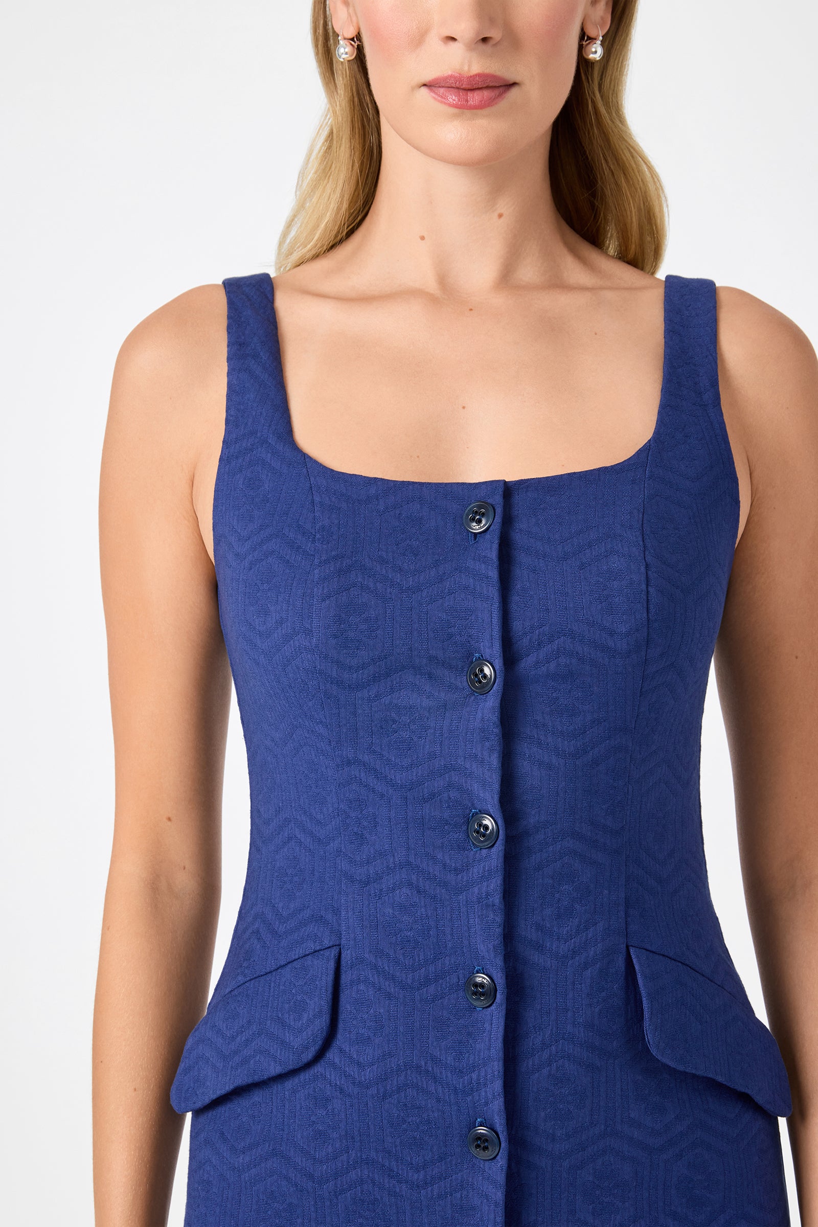 A woman wears the Shinra Button-Down Mini Dress—a sleeveless, textured blue dress with a square neckline, front buttons, and two flap pockets—shown from shoulders to hips against a plain background.