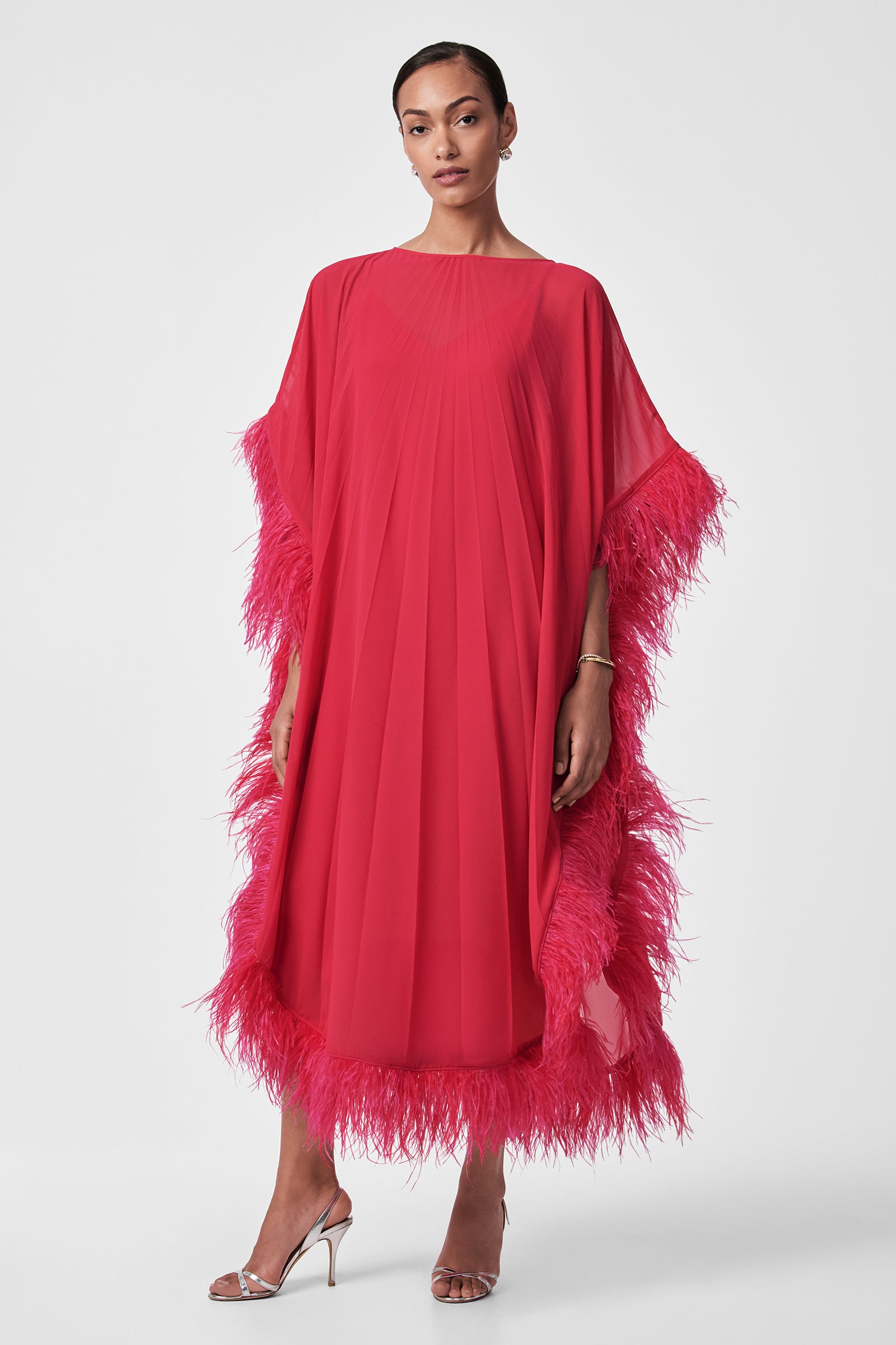 A woman poses against a white background in the Pleated Neena Feather-Trimmed Caftan Dress, a bright pink, loose-fitting style with pleats and feathered trim on the sleeves, hem, and sides. She pairs it with silver high-heeled sandals and wears her hair pulled back.