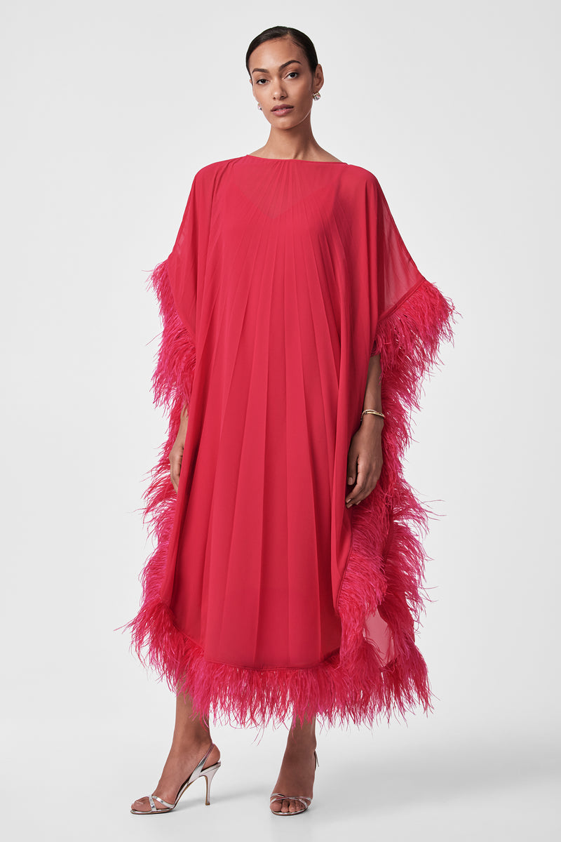 A woman poses against a white background in the Pleated Neena Feather-Trimmed Caftan Dress, a bright pink, loose-fitting style with pleats and feathered trim on the sleeves, hem, and sides. She pairs it with silver high-heeled sandals and wears her hair pulled back.