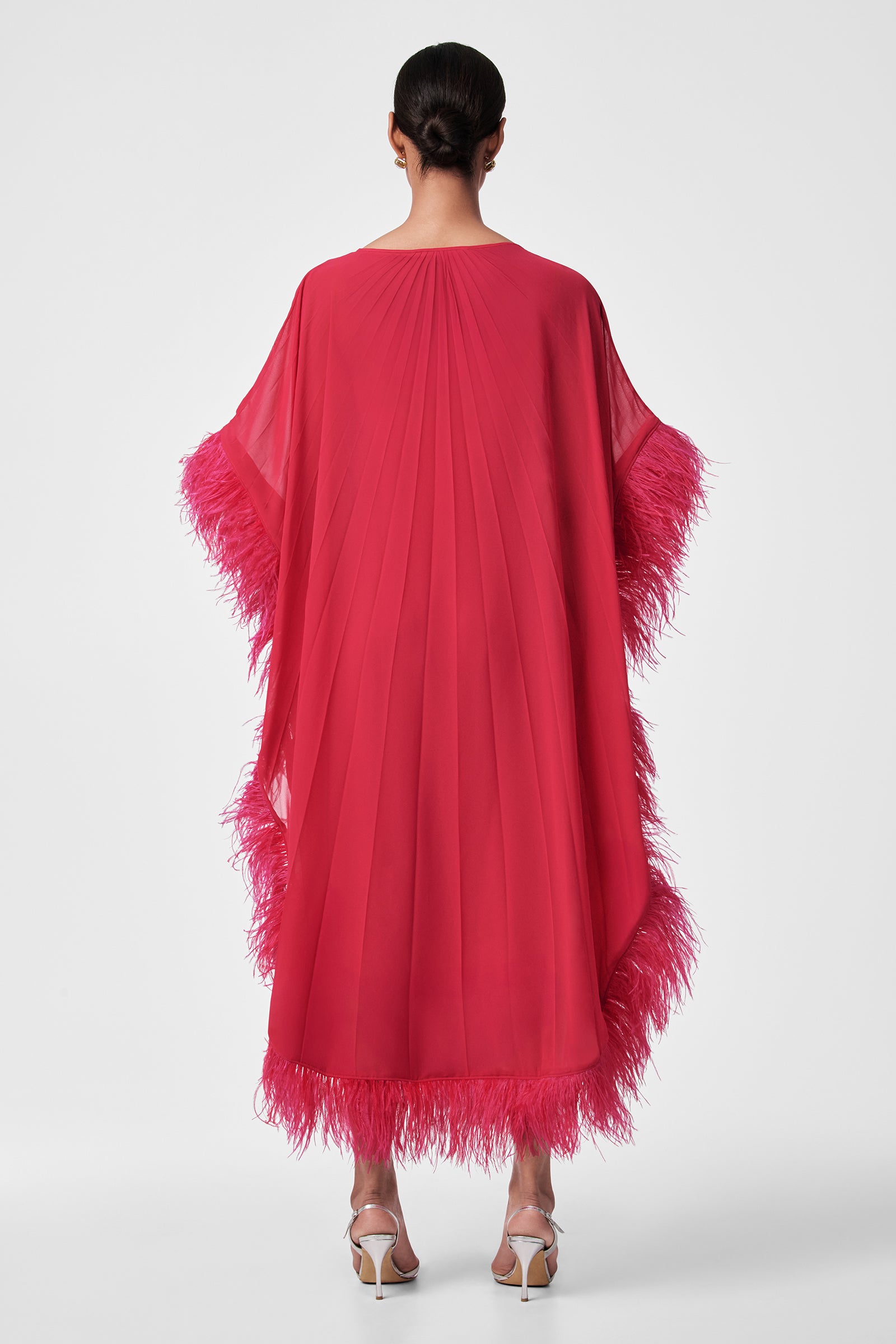 A woman faces away, wearing the Pleated Neena Feather-Trimmed Caftan Dress in bright pink with pleats and feather trim on the sleeves and hem. Her hair is styled in a low bun, paired with white open-heeled sandals against a plain white background.