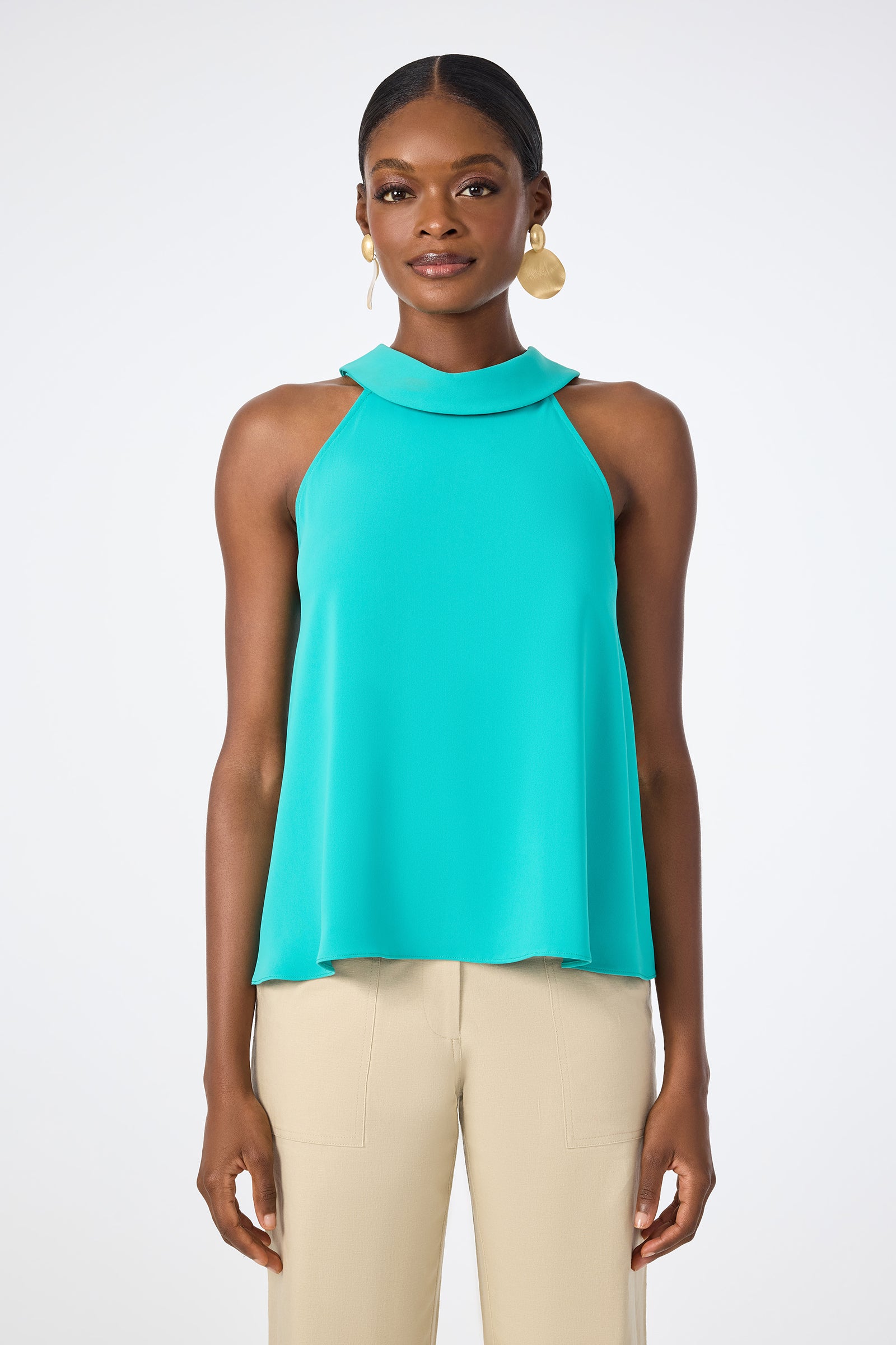 A woman stands against a white background wearing the Eze Halter Top in sleeveless turquoise with a wide collar, paired with beige pants. She accessorizes with gold hoop earrings and has her hair pulled back.