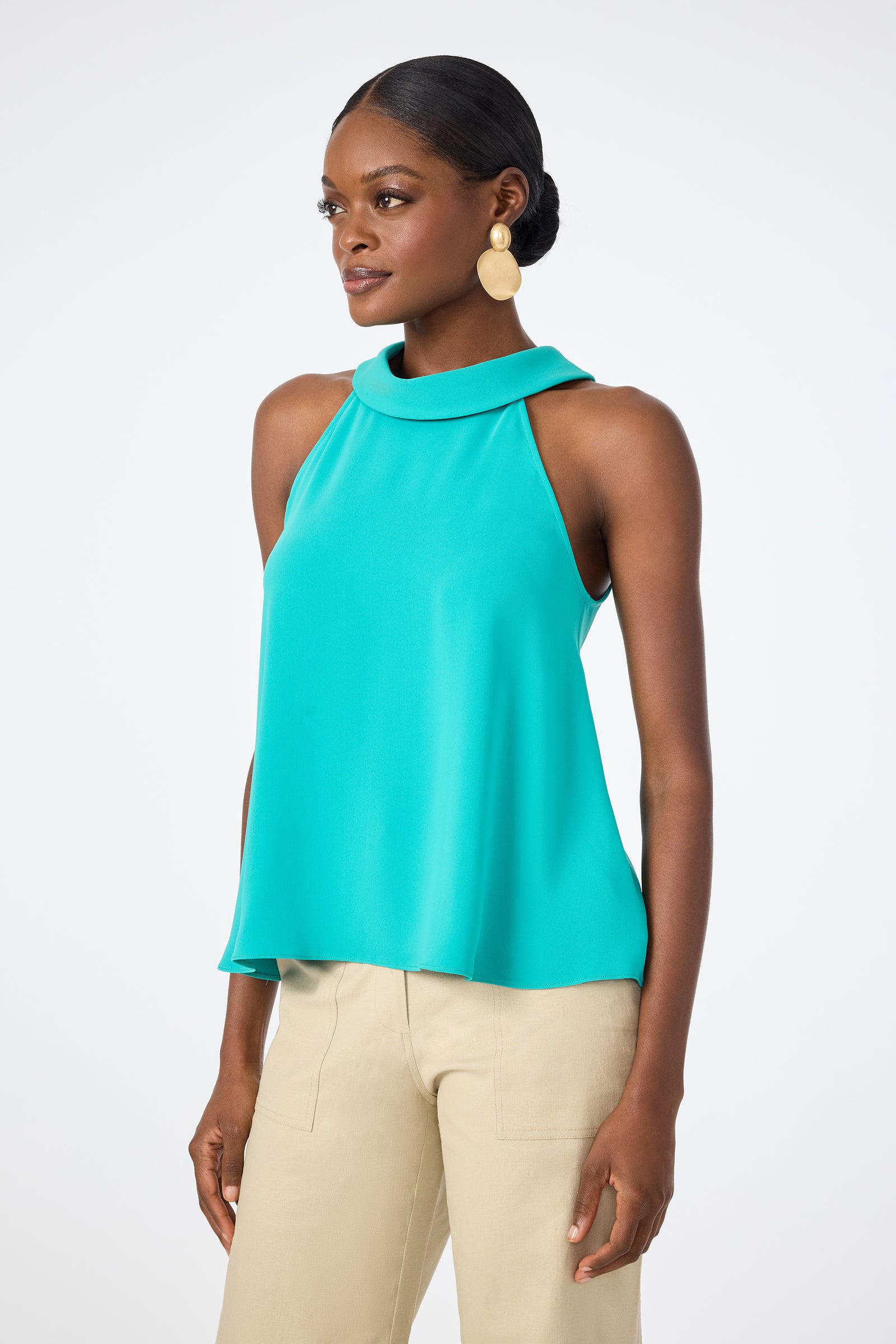A woman wears the Eze Halter Top in turquoise with beige pants, her hair pulled back and accessorized with large round gold earrings. She stands slightly turned to the side against a plain white background.