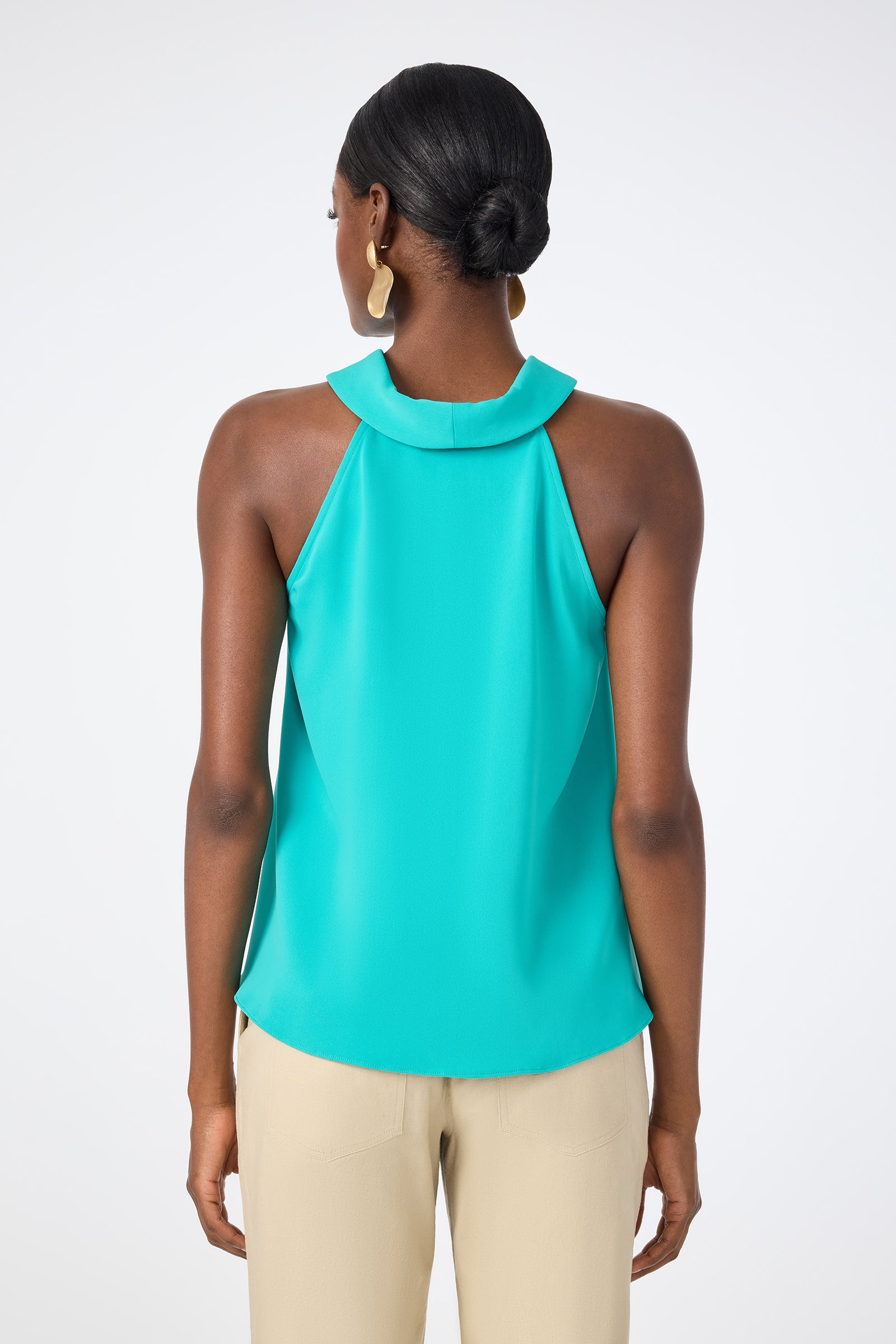 A woman with dark hair in a low bun wears the sleeveless Eze Halter Top in turquoise and light beige pants, standing with her back to the camera against a plain white background.