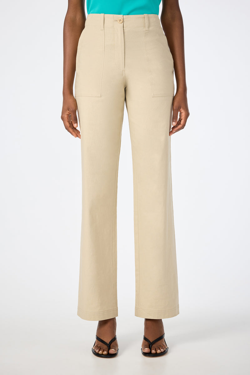 Someone stands against a plain white background wearing Antonia 2 Straight Leg Pants in Biarritz Beige with front patch pockets, paired with a turquoise top and open-toe sandals.