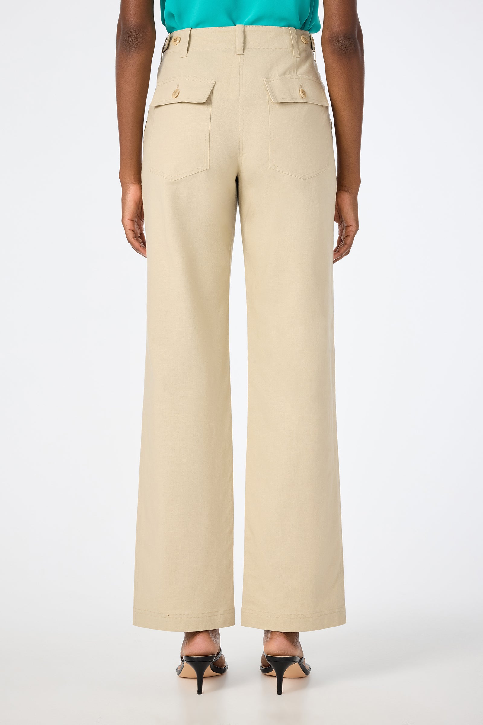 A person wearing Antonia 2 Straight Leg Pants in beige, featuring high waist, wide legs, back flap pockets, and tan buttons, stands in heels with their back to a plain white background.