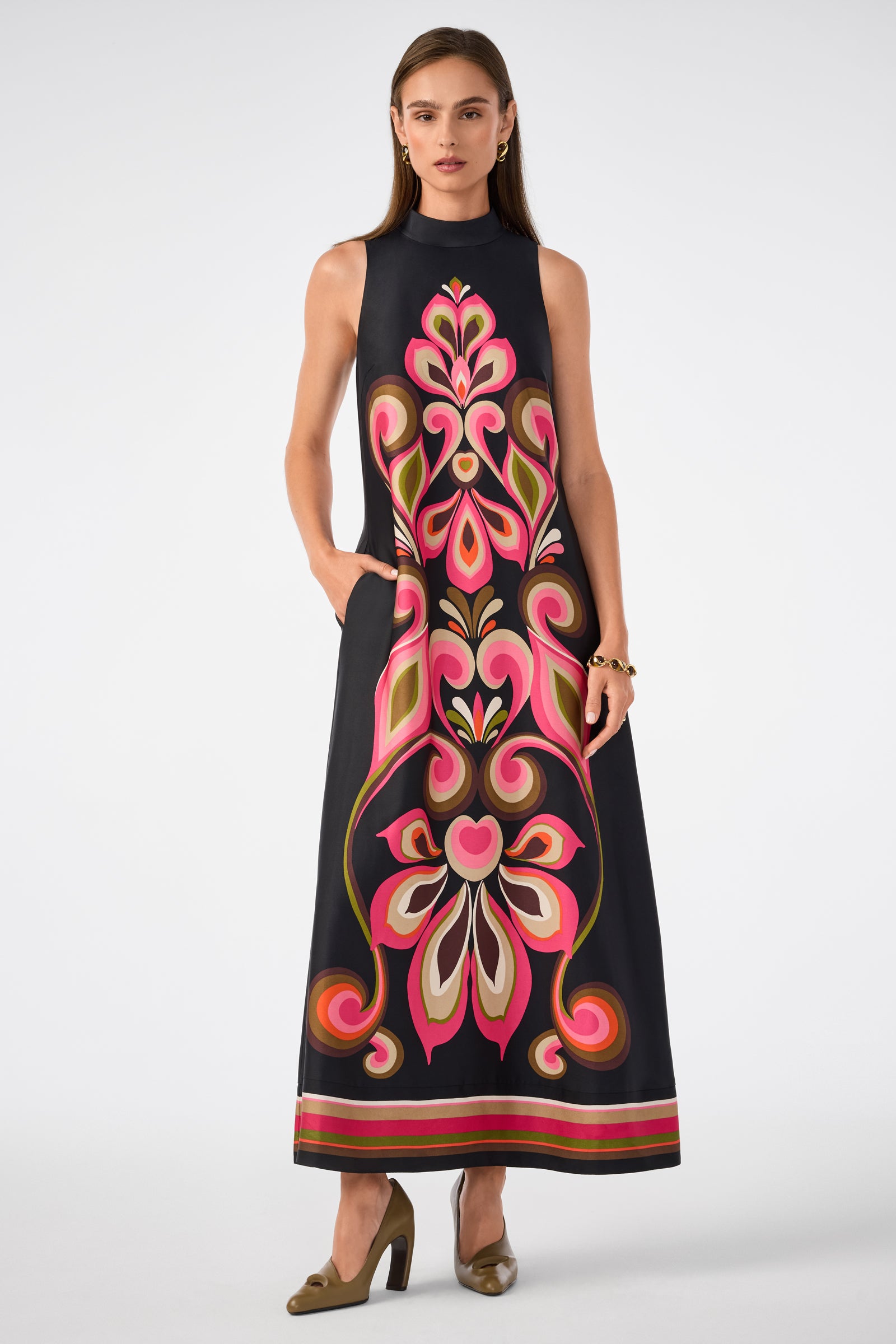 A woman wears the Loveage Sleeveless Maxi Dress, a floor-length black dress with a bold, colorful floral pattern in pink, red, green, and beige tones, paired with olive green heels.
