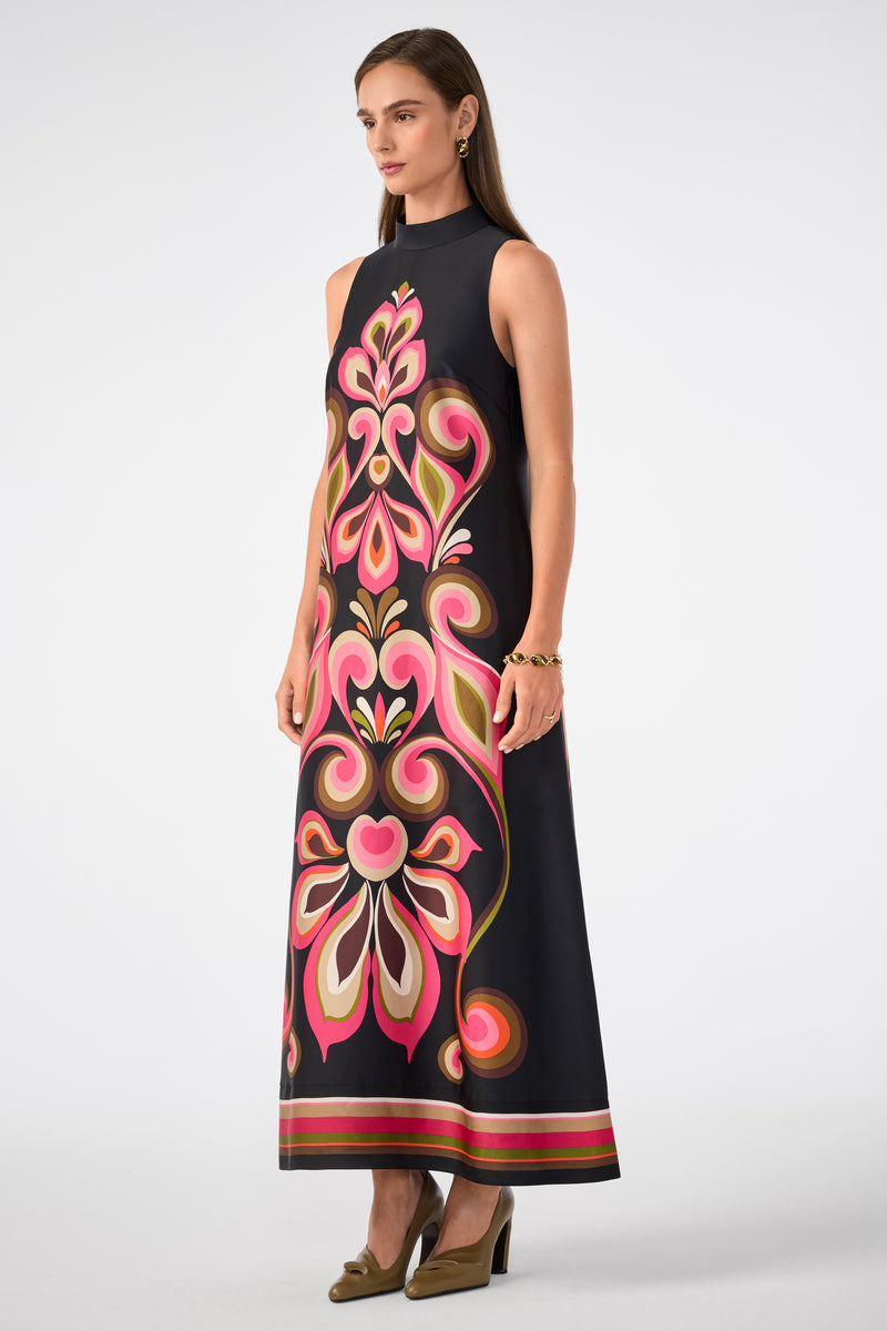 A woman models the Loveage Sleeveless Maxi Dress, a floor-length black dress with bold pink, red, beige, and orange floral patterns. She pairs it with olive green heels, gold jewelry, and wears her brown hair straight.