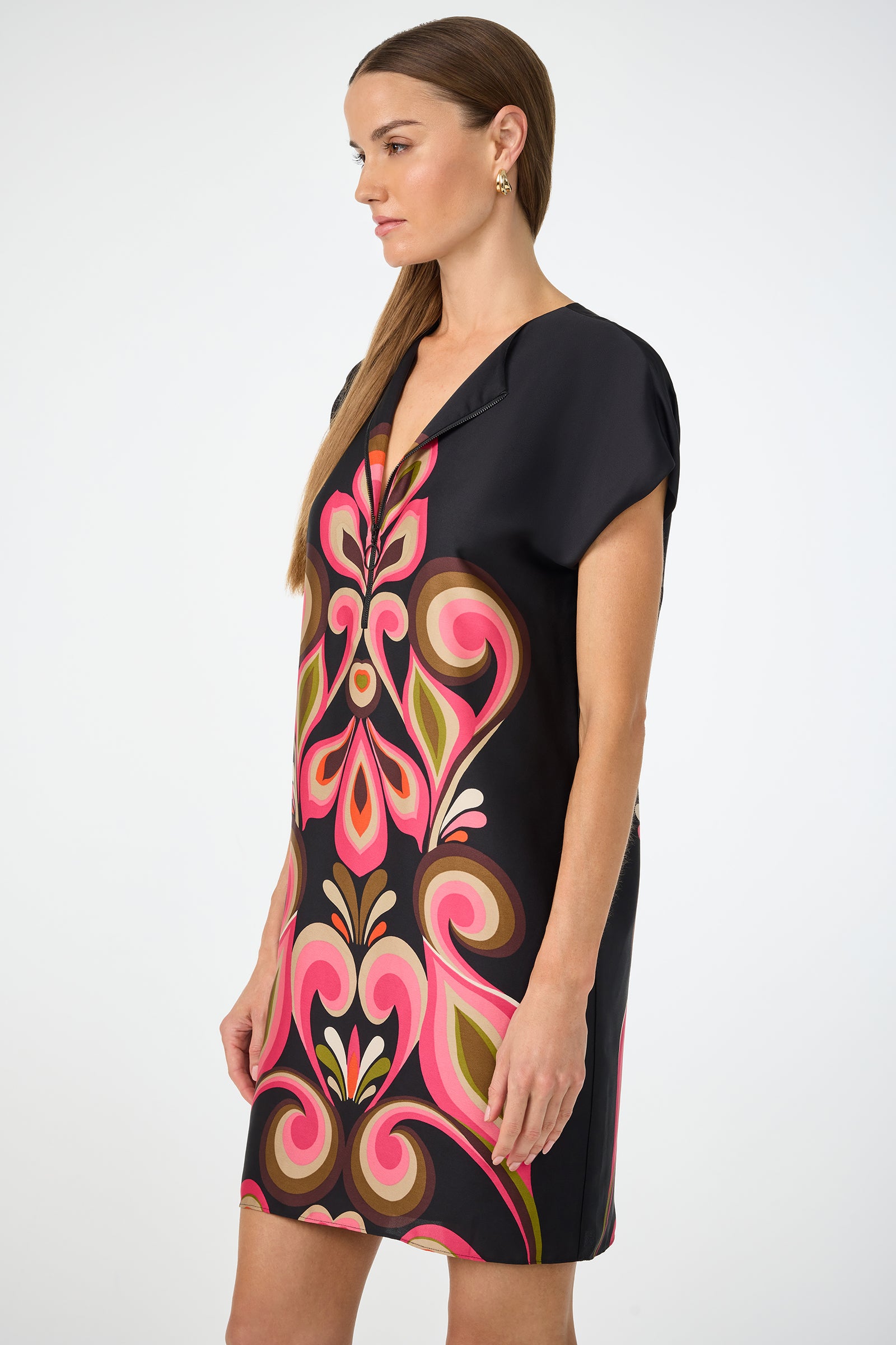 A woman in profile wears the Hasil 3 Short-Sleeve Shift Dress by Sanctuary, featuring a bold black base with vibrant pink, orange, and green abstract patterns. She has straight brown hair and accessorizes with small gold earrings.