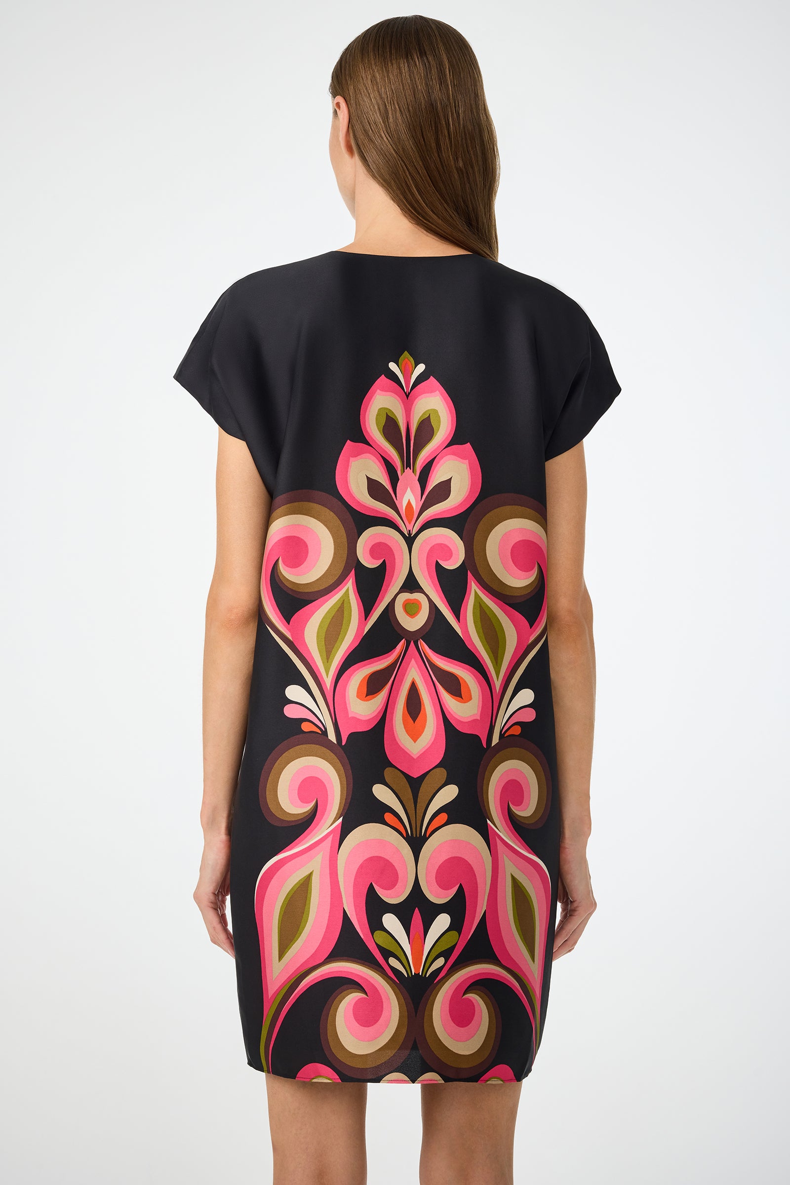A woman with straight brown hair is seen from behind wearing the Hasil 3 Short-Sleeve Shift Dress by Sanctuary in black, featuring a vibrant pink, green, and brown symmetrical floral pattern on the back against a white background.