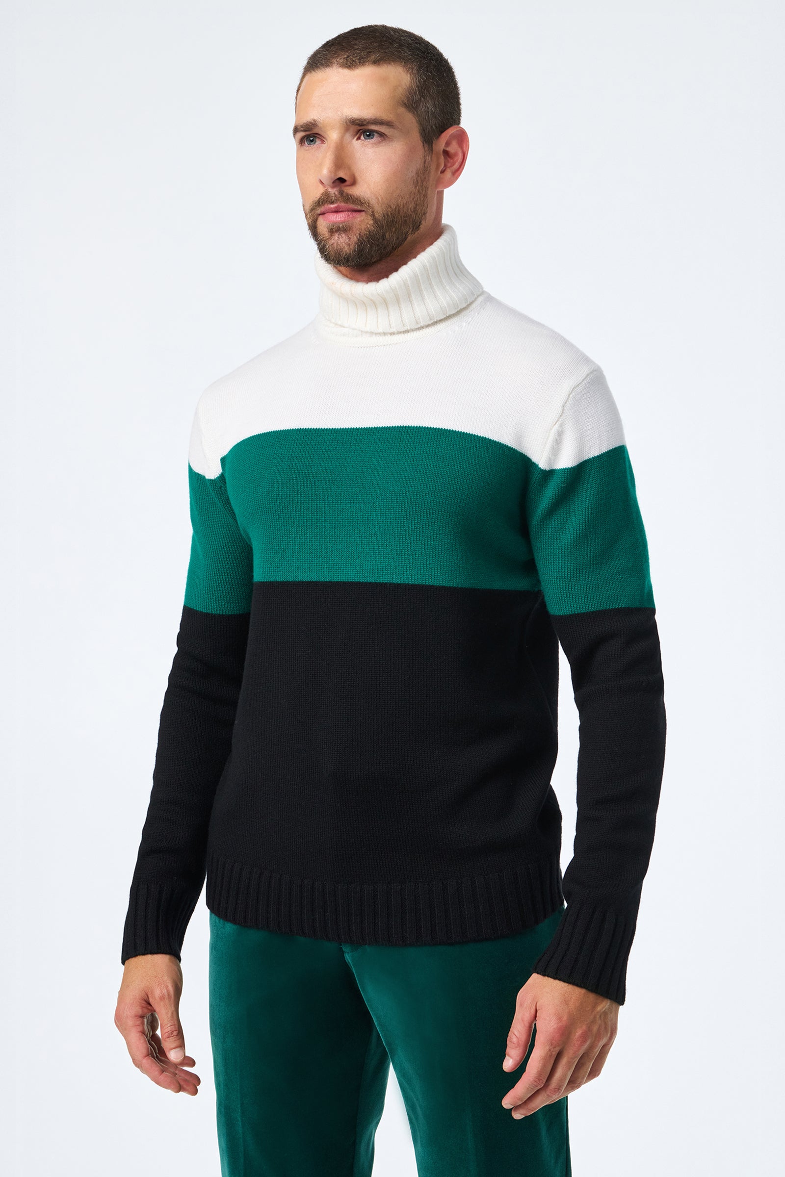 A man with short hair and a beard wears the Harrelson T-Neck color-block sweater in white, green, and black, paired with green velvet pants, standing against a plain white background.